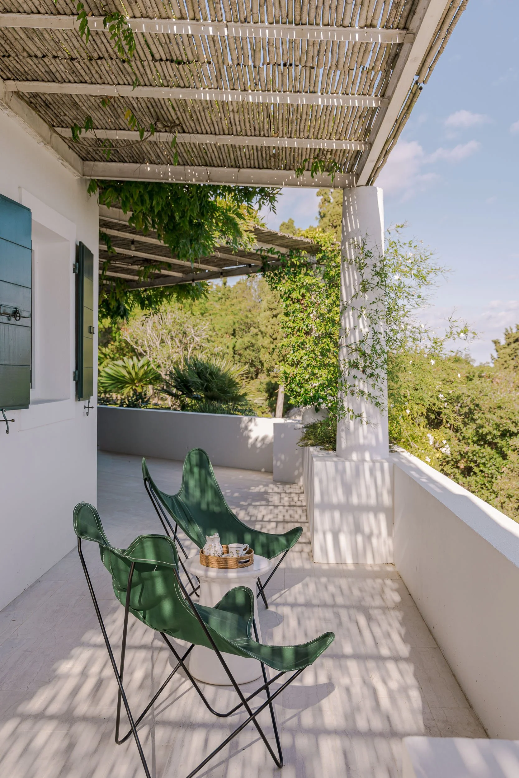 A cozy outdoor balcony with three green butterfly chairs around a white side table holding a tray with a teapot and cups, shaded by a bamboo pergola with vines, overlooking lush green trees.