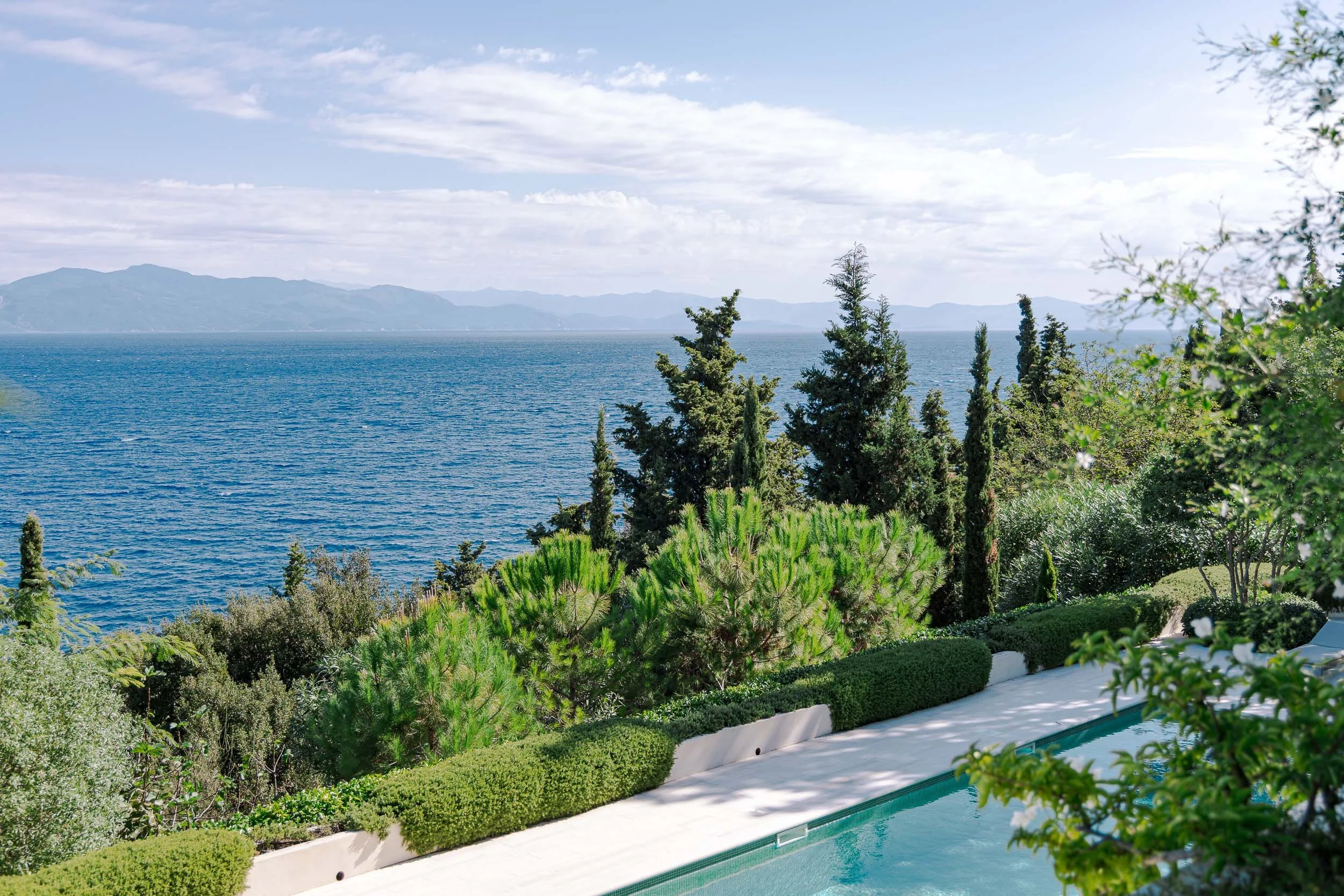 View of the ocean with mountains in the distance, with lush green trees and bushes along the edge of a swimming pool in the foreground.