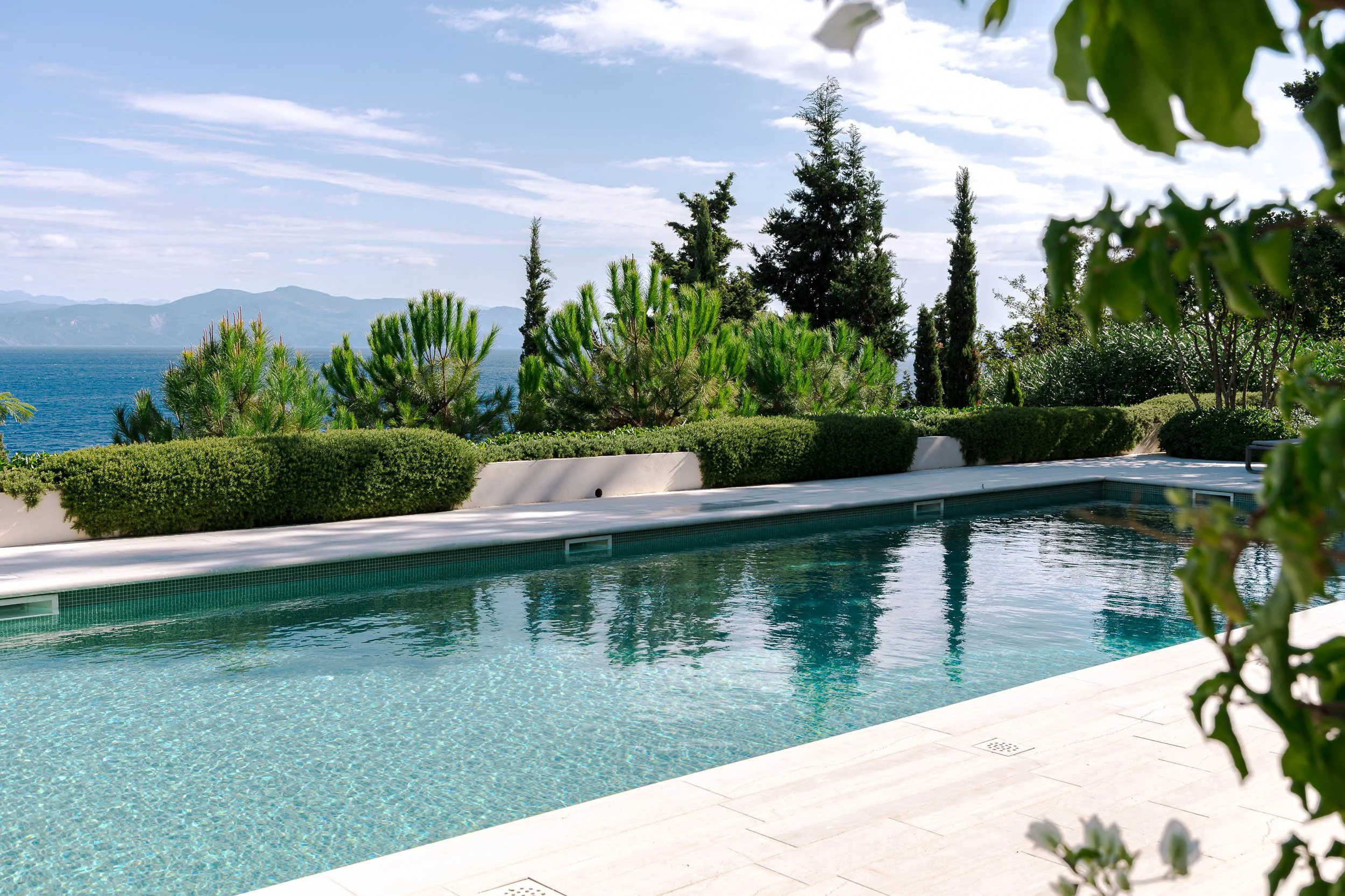 View of a swimming pool with a white border, surrounded by greenery, including shrubs and trees, with a scenic view of the ocean and mountains in the background under a partly cloudy sky.