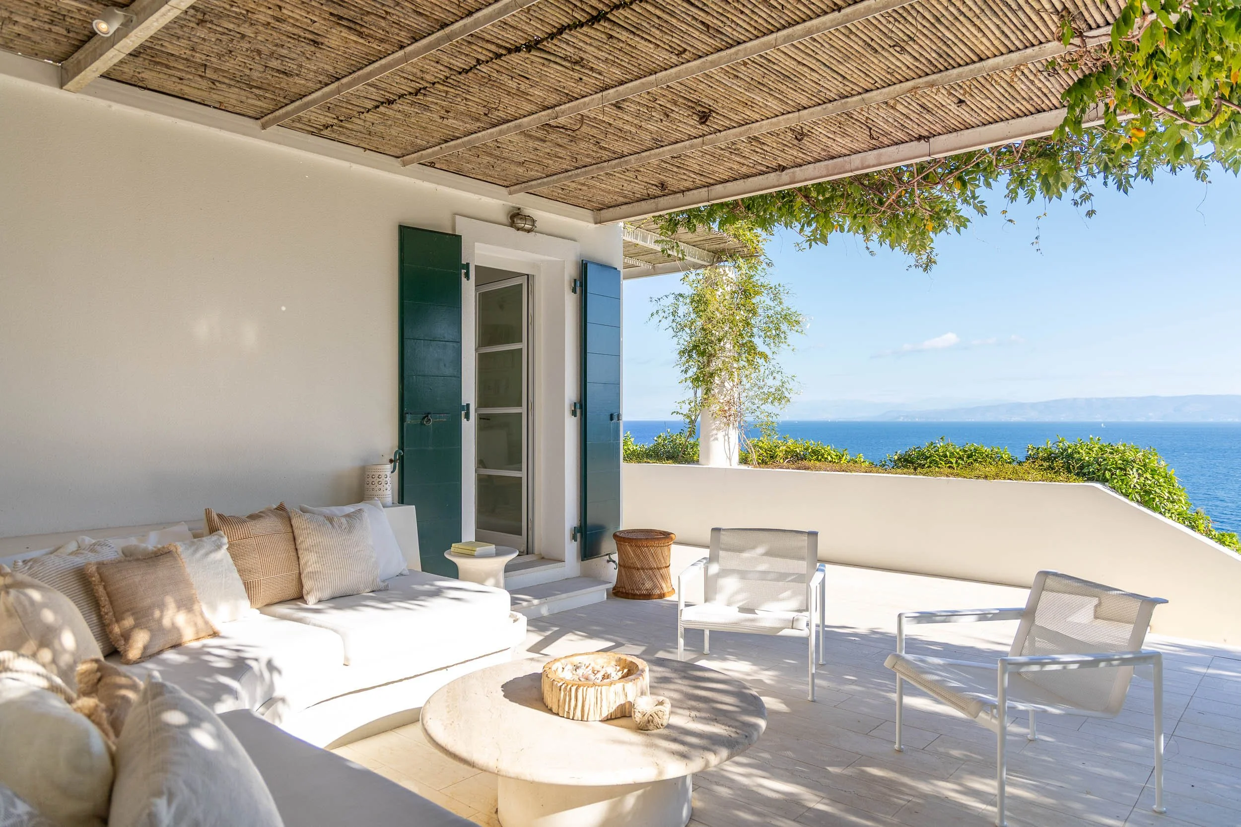 A balcony overlooking the ocean with white walls and furniture, blue shutters, a white sofa with neutral cushions, a round stone coffee table, two white outdoor chairs, and greenery, with a view of the sea and sky in the background.
