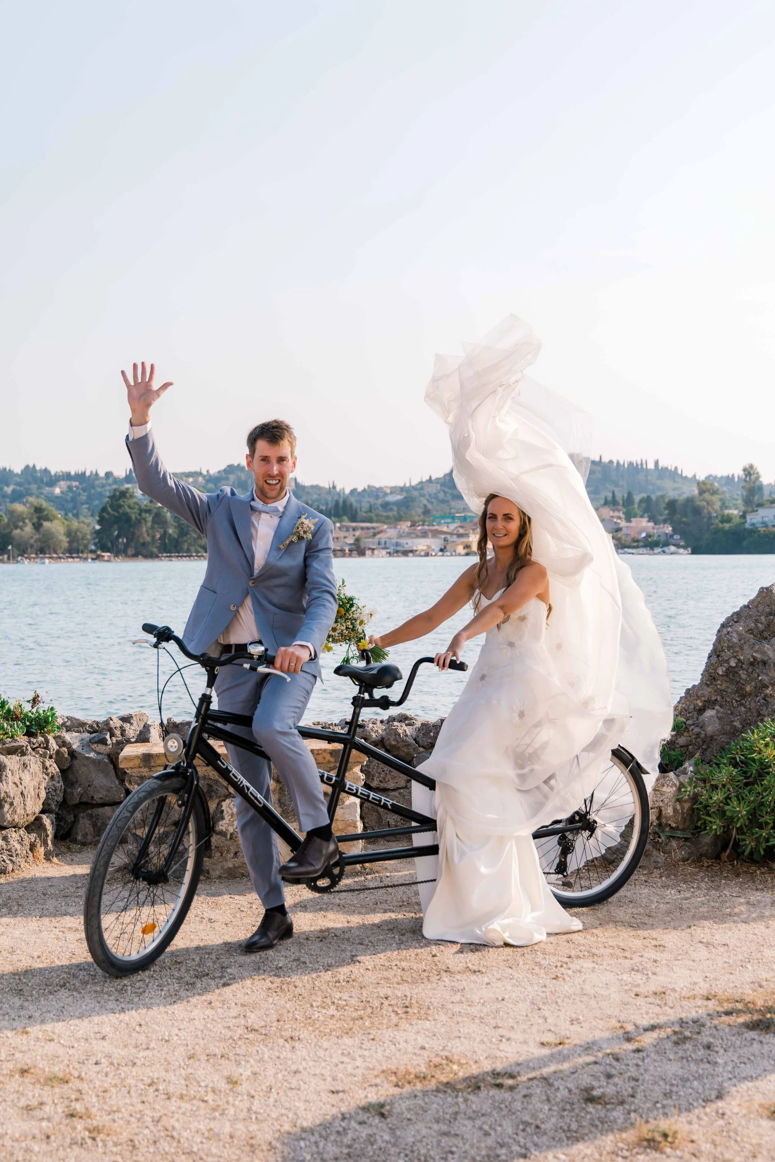 A man in a gray suit and a woman in a white wedding dress riding a tandem bicycle along a waterfront. The man is waving, and the woman is holding the handlebars, with her wedding dress flowing. There are trees, water, and hills in the background.