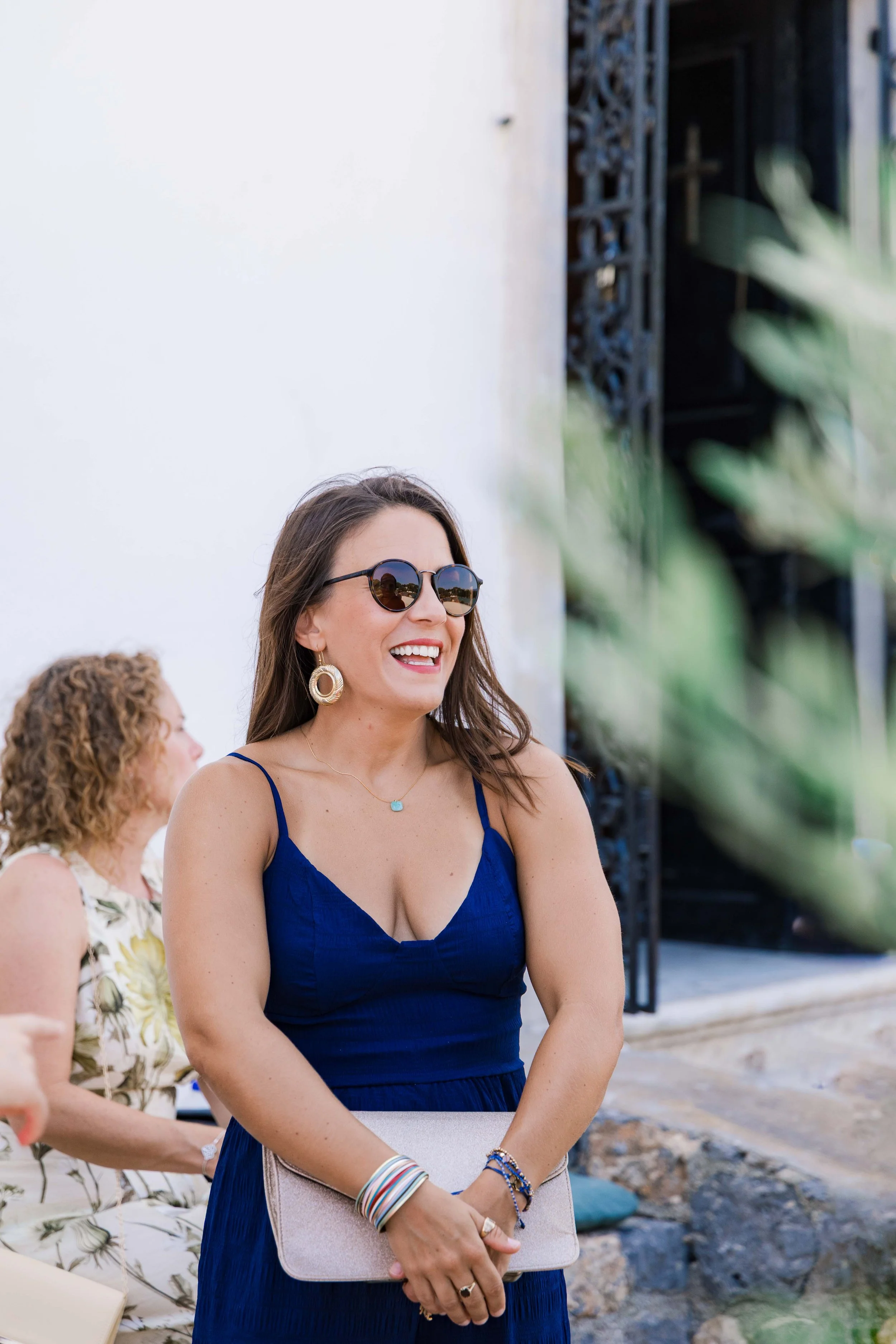A woman with long brown hair wearing black sunglasses, a blue dress with spaghetti straps, gold earrings and bracelets, holding a beige clutch, smiling at an outdoor event. There are other women in the background and a stone sidewalk with greenery.