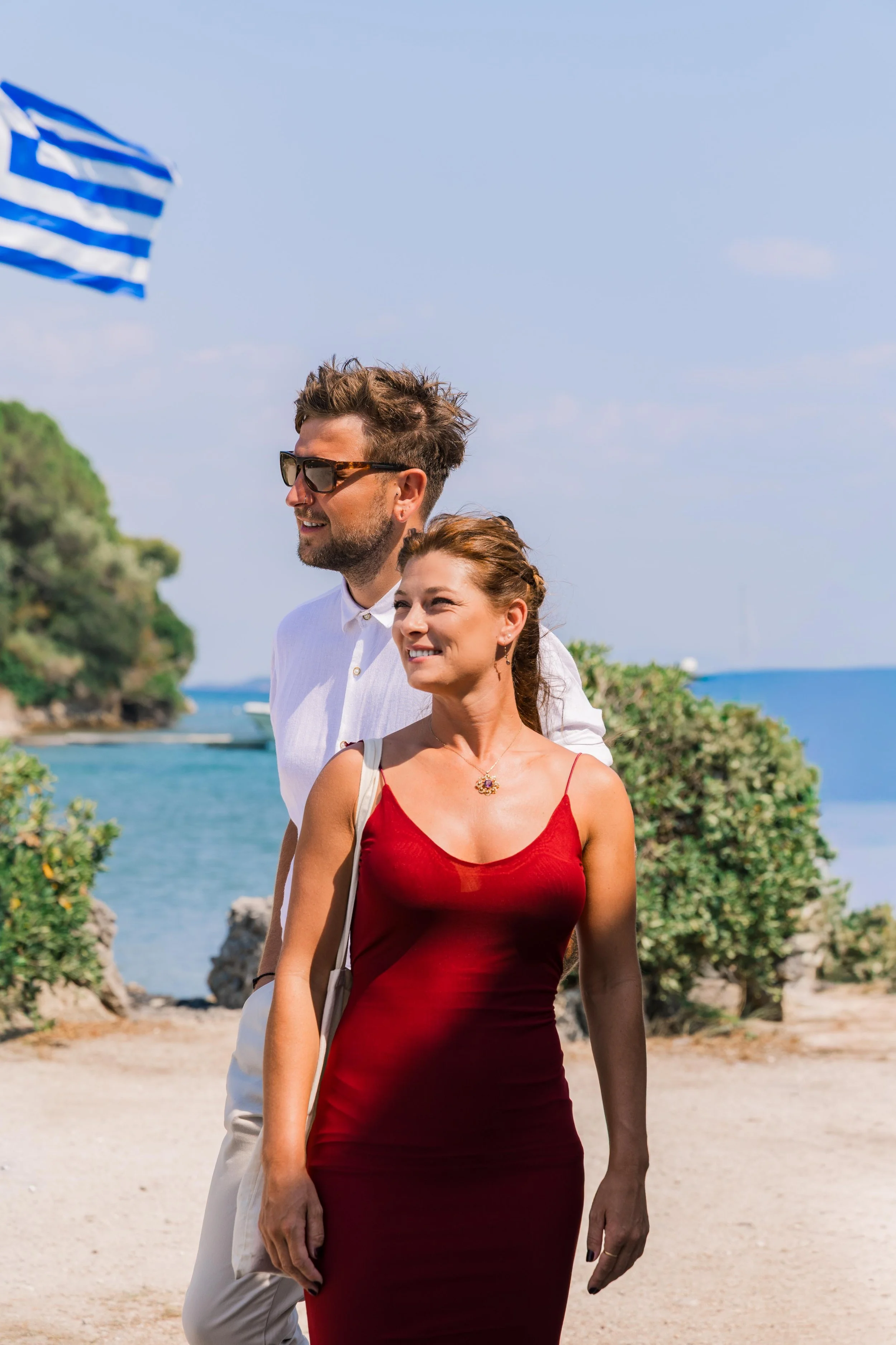 A man and woman walking together outdoors near the coast. The woman is wearing a red dress and a necklace, and the man is wearing a white shirt, beige pants, and sunglasses. There is a Greek flag in the background, along with greenery and the sea.
