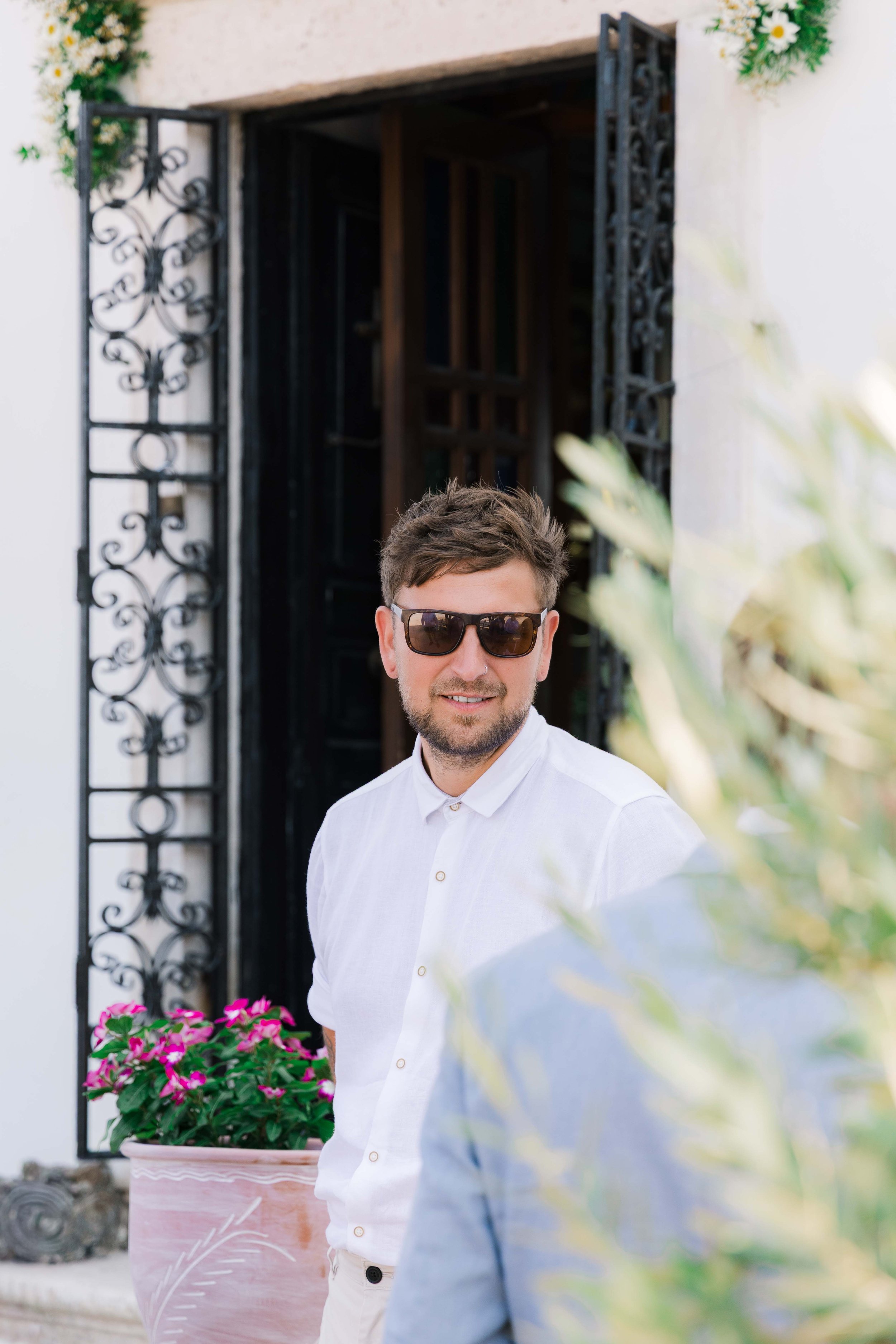 Young man with brown hair and beard wearing sunglasses and a white shirt, standing outdoors near a building with ornate ironwork on the window frame, pink flowers in a pot, and greenery.