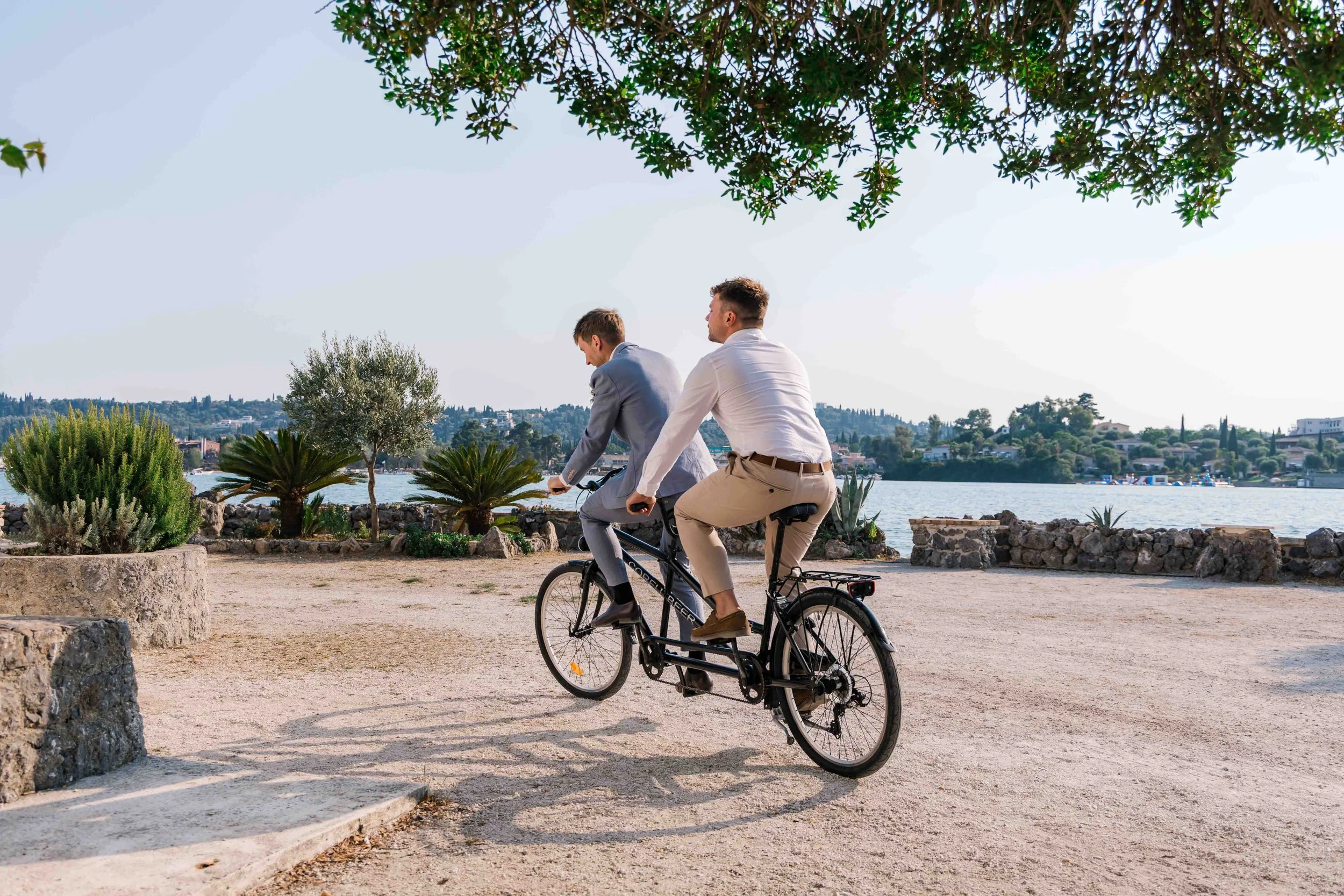 Two men in business attire riding a tandem bicycle along a waterfront park, with green plants, rocky border, and water in the background during daylight.