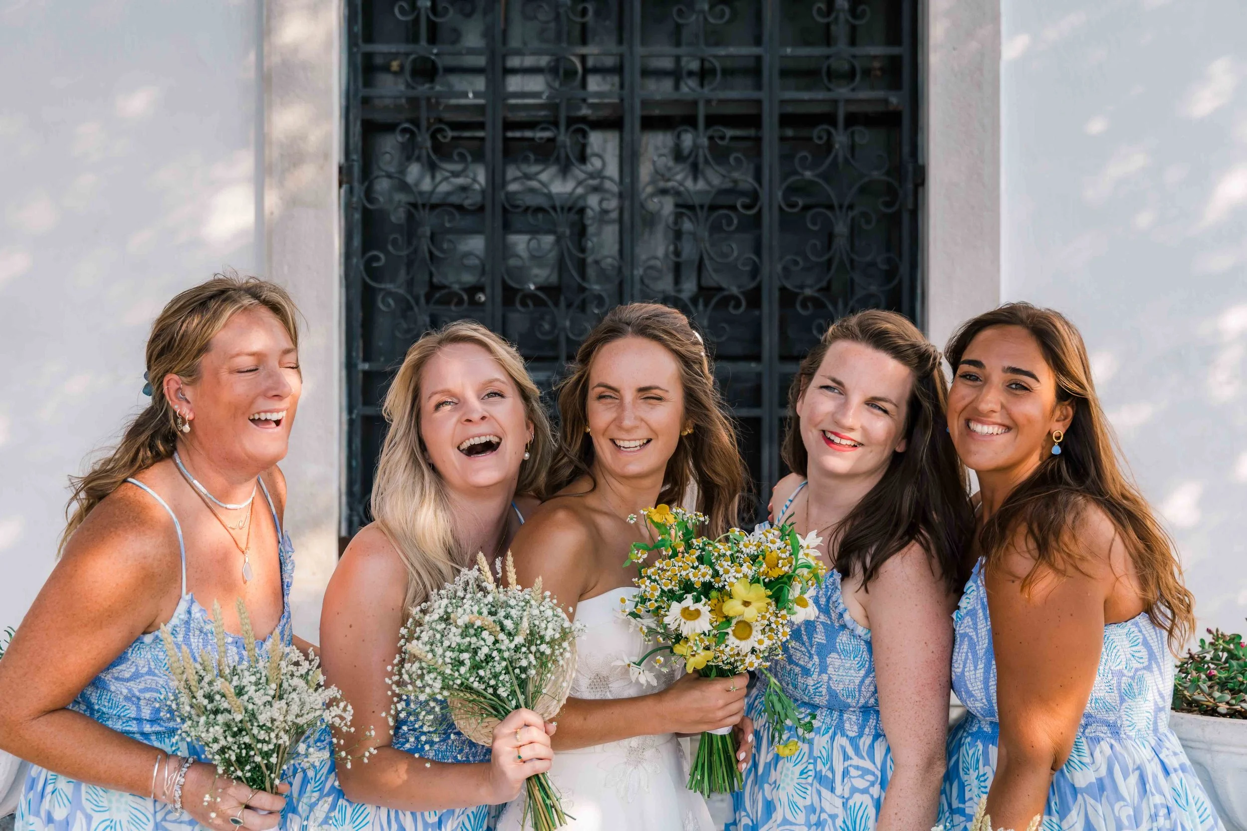 A group of six women gathered together outside in front of a metal gate, smiling and holding bouquets of flowers, dressed in matching blue and white patterned dresses, celebrating a special occasion.