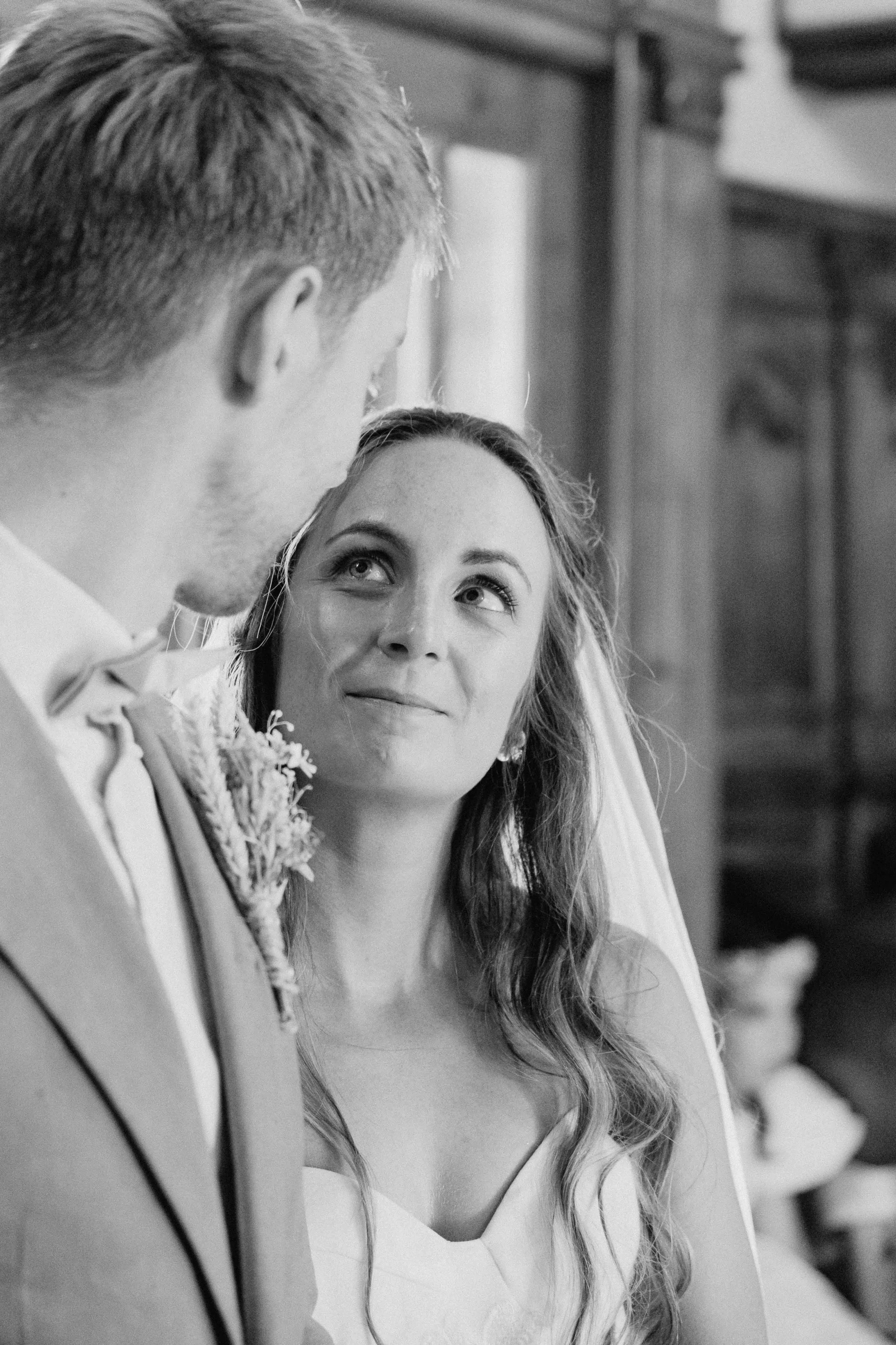 A bride with curly hair and makeup looking up at a groom with short hair and a tuxedo, in a wedding setting.