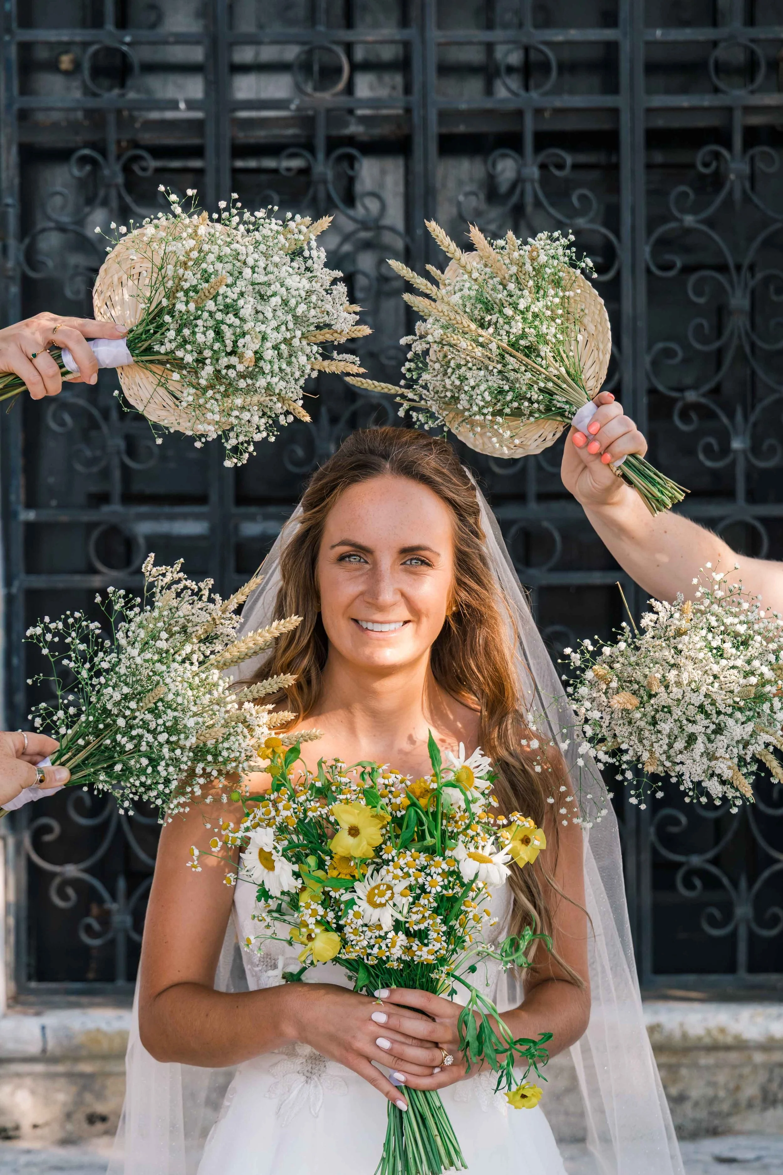 A smiling bride in a wedding dress holding a bouquet of yellow and white flowers, with two hands holding smaller bouquets of white flowers above her head against a black wrought iron gate background.