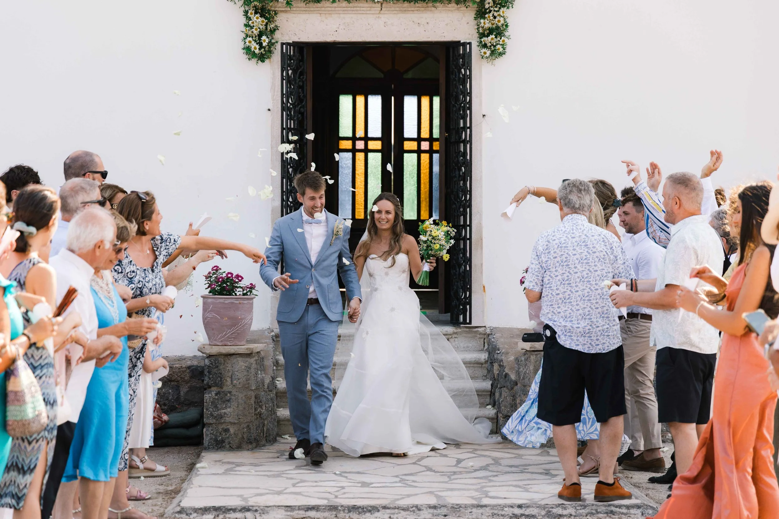 A bride and groom leaving a church while guests throw flower petals at them. The couple is smiling and holding hands, with the bride holding a bouquet of flowers, as they walk down the steps.