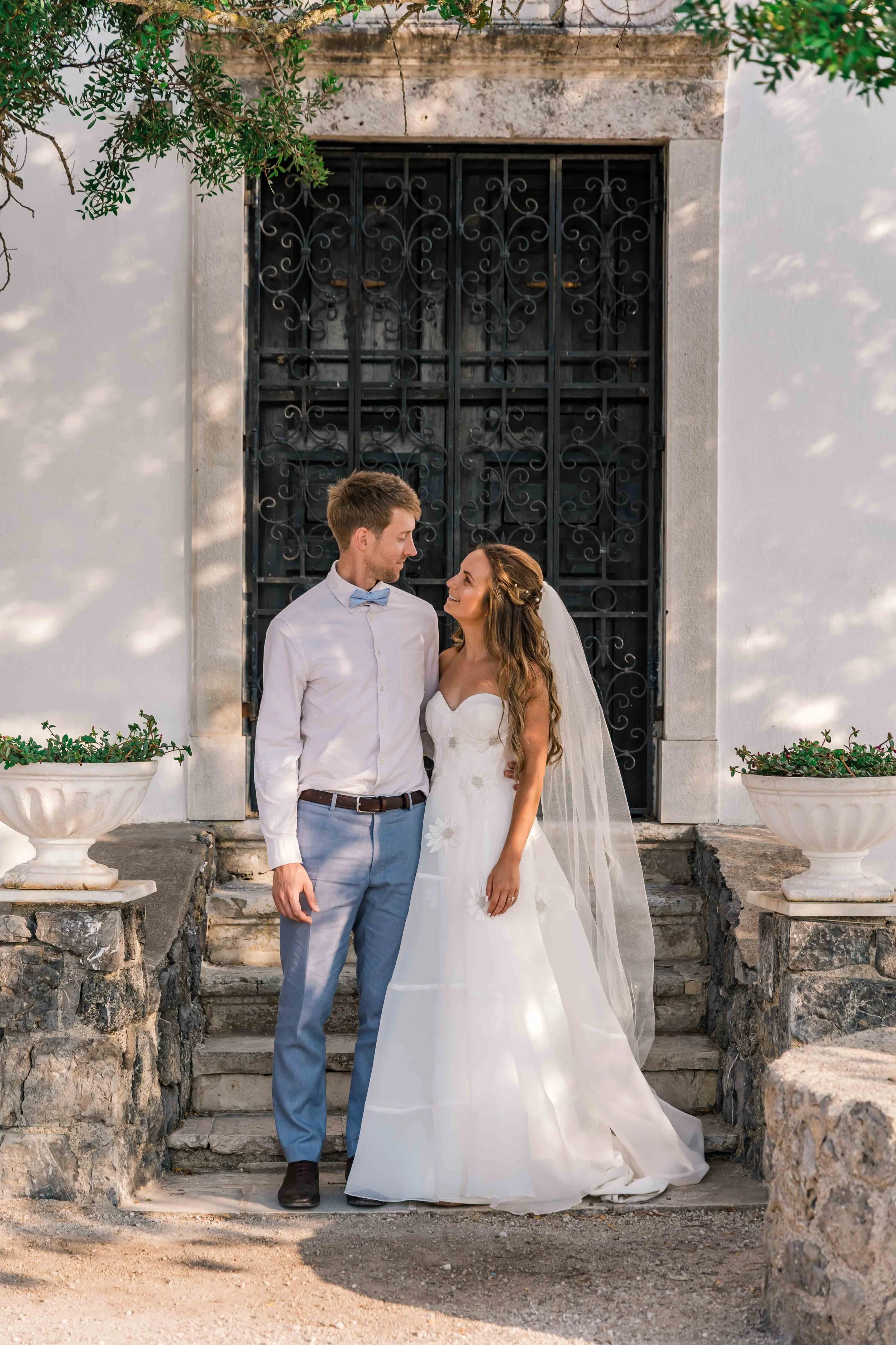 A bride and groom standing on stone stairs outside a building with black wrought iron doors. The bride is wearing a white strapless wedding gown and veil, and the groom is in a light blue suit with a bow tie. They are gazing at each other affectionat