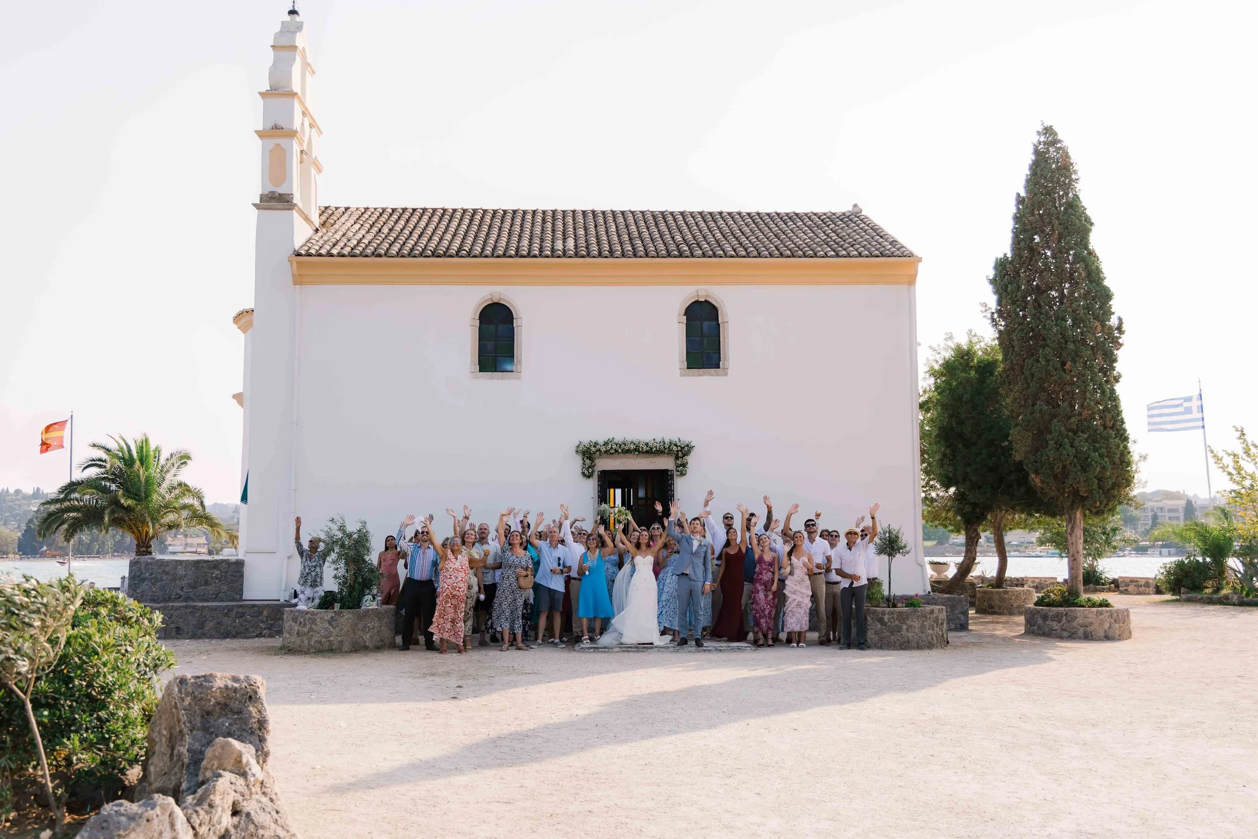 A group of people gathered outside a white church with arched windows, raising their hands in celebration, with palm trees and flags in the background.
