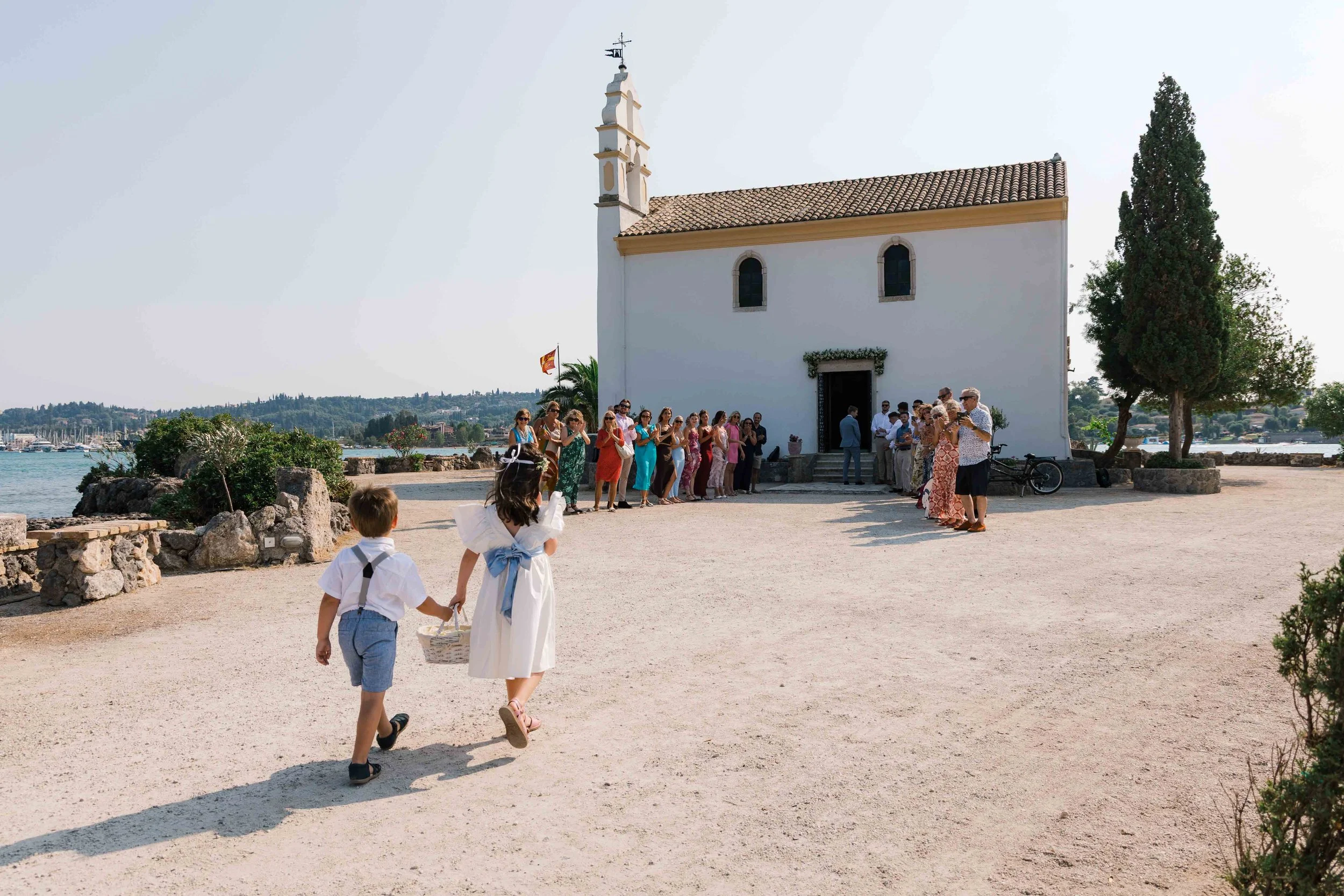 Children dressed in white with blue sashes walk hand in hand toward a group of people gathered outside a white church by the water. The group appears to be preparing for a ceremony.