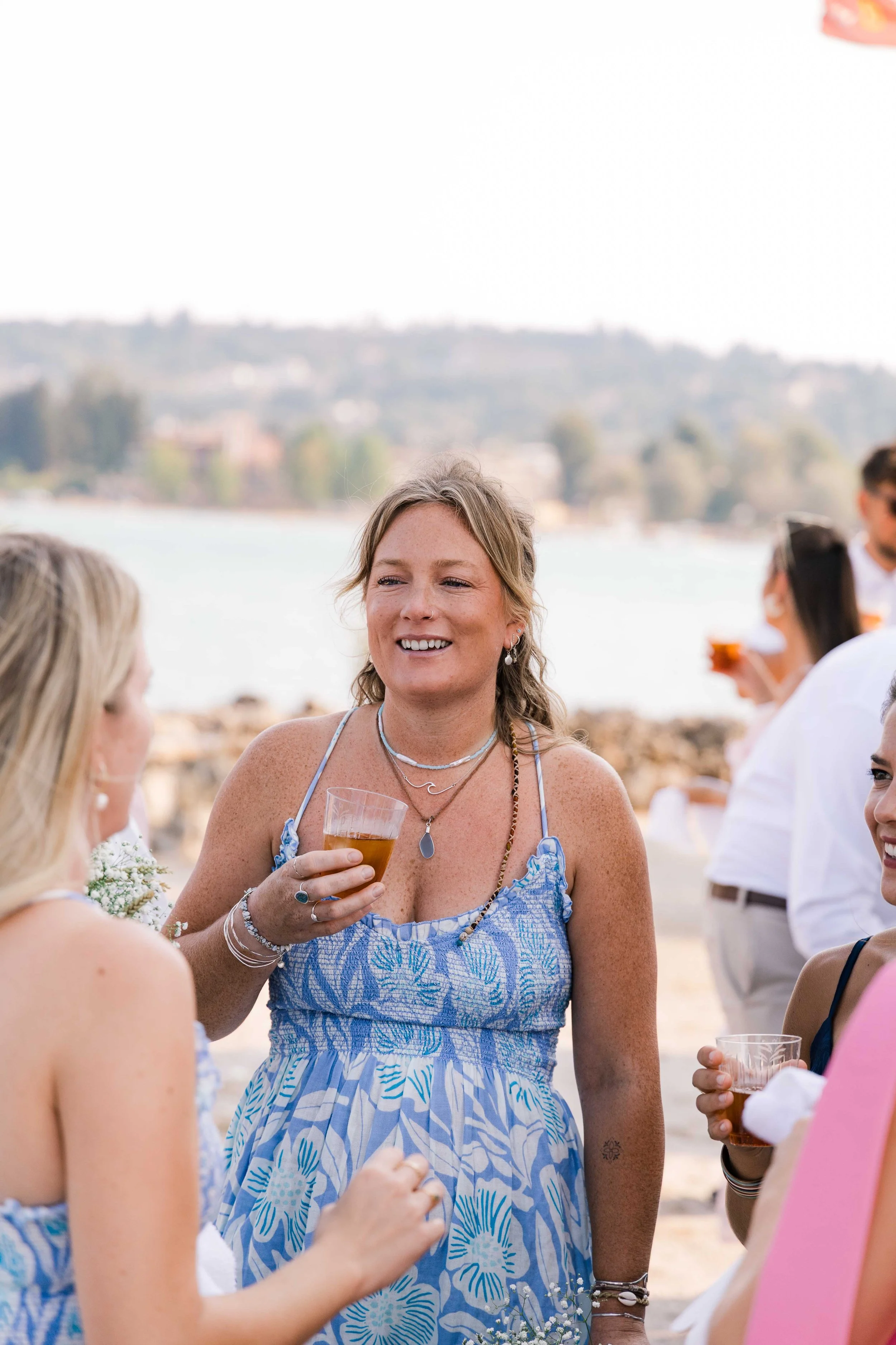A woman with blonde hair holding a drink at an outdoor gathering by a body of water, smiling and engaging with others, wearing a blue floral dress.