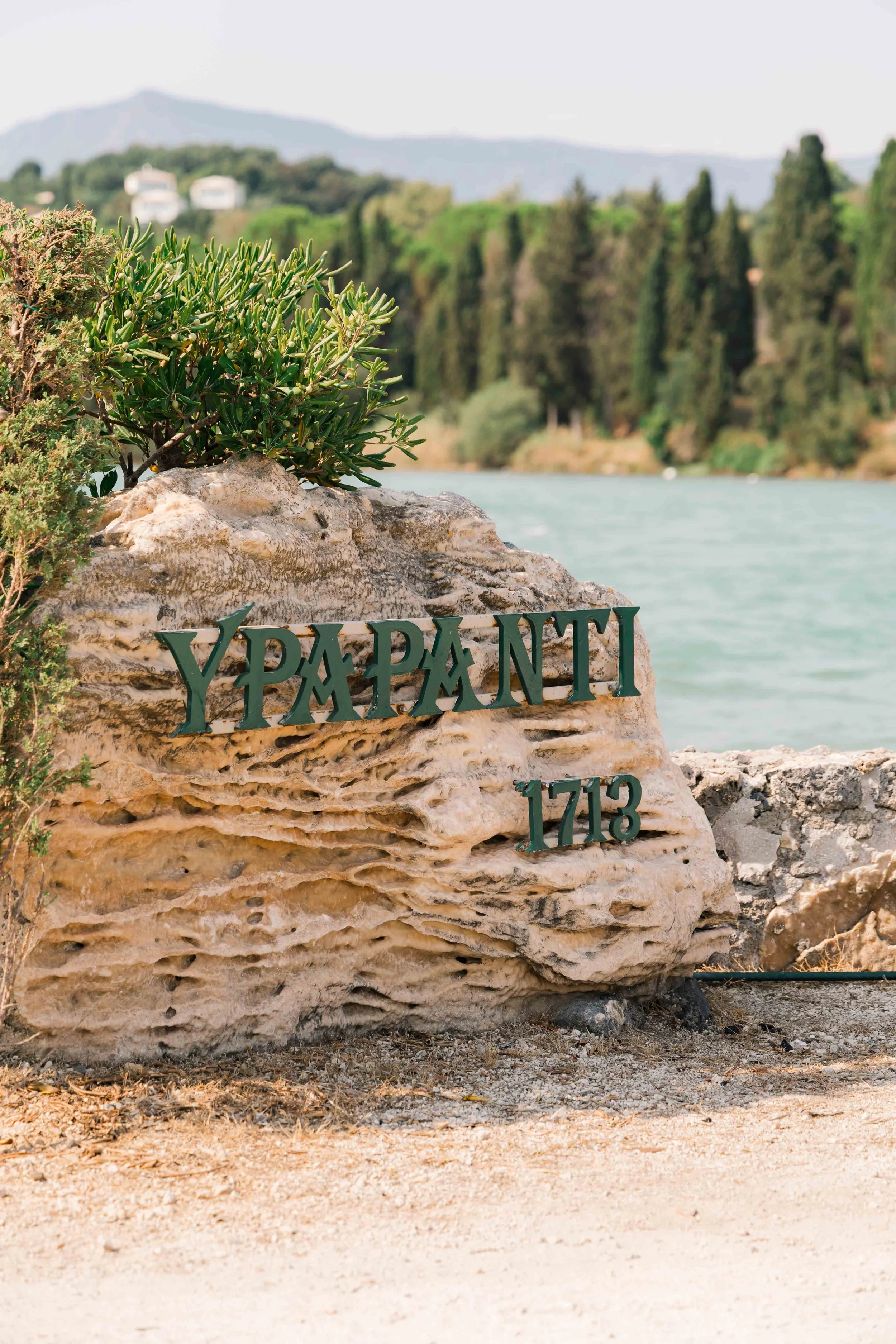 A large rock with the word 'Y Phoenix' and the number '1713' on it, surrounded by small plants, with a body of water and green trees in the background.