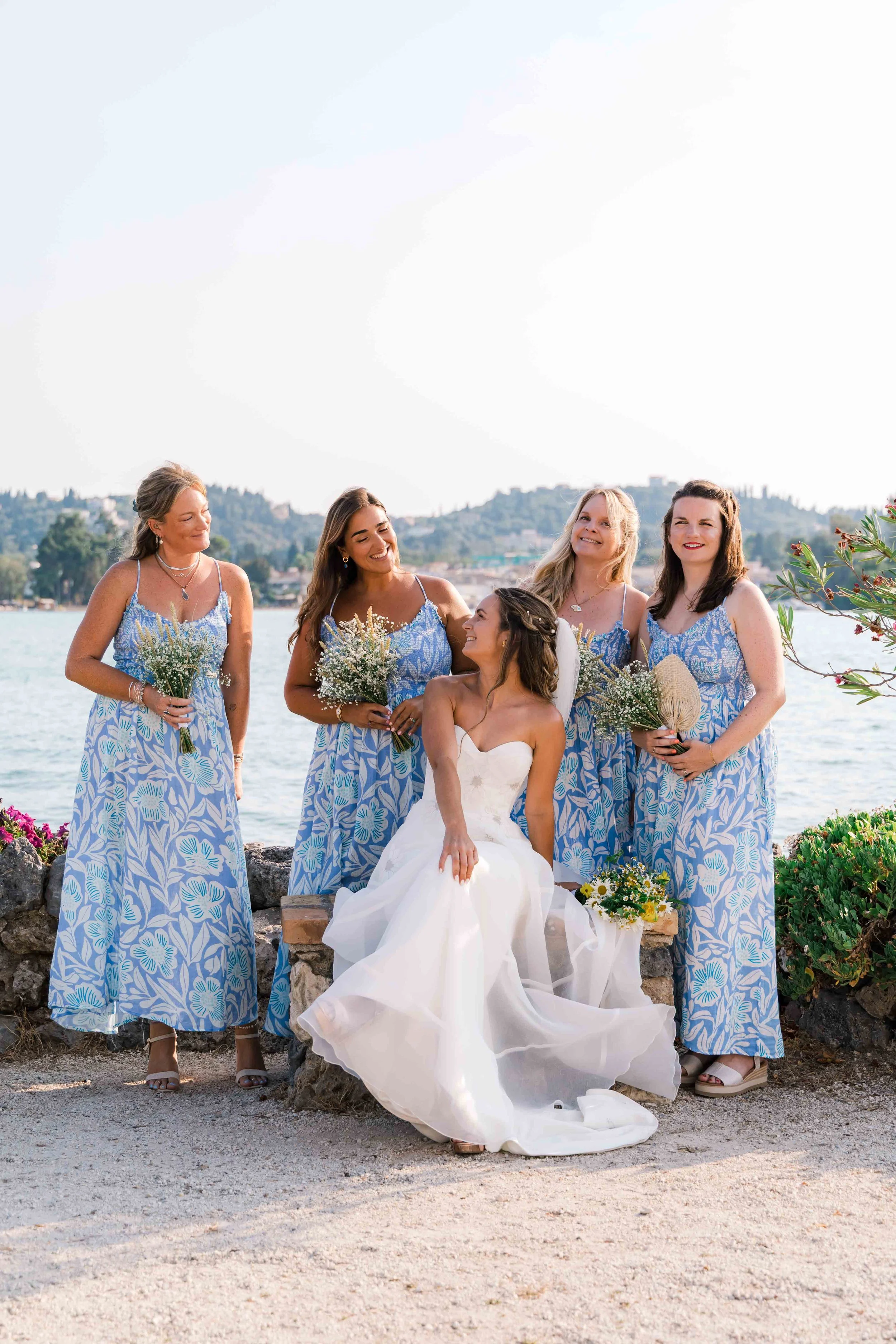 Group of five women in matching blue floral dresses, one woman in a white wedding gown, at a beachside wedding ceremony with a body of water and hills in the background.