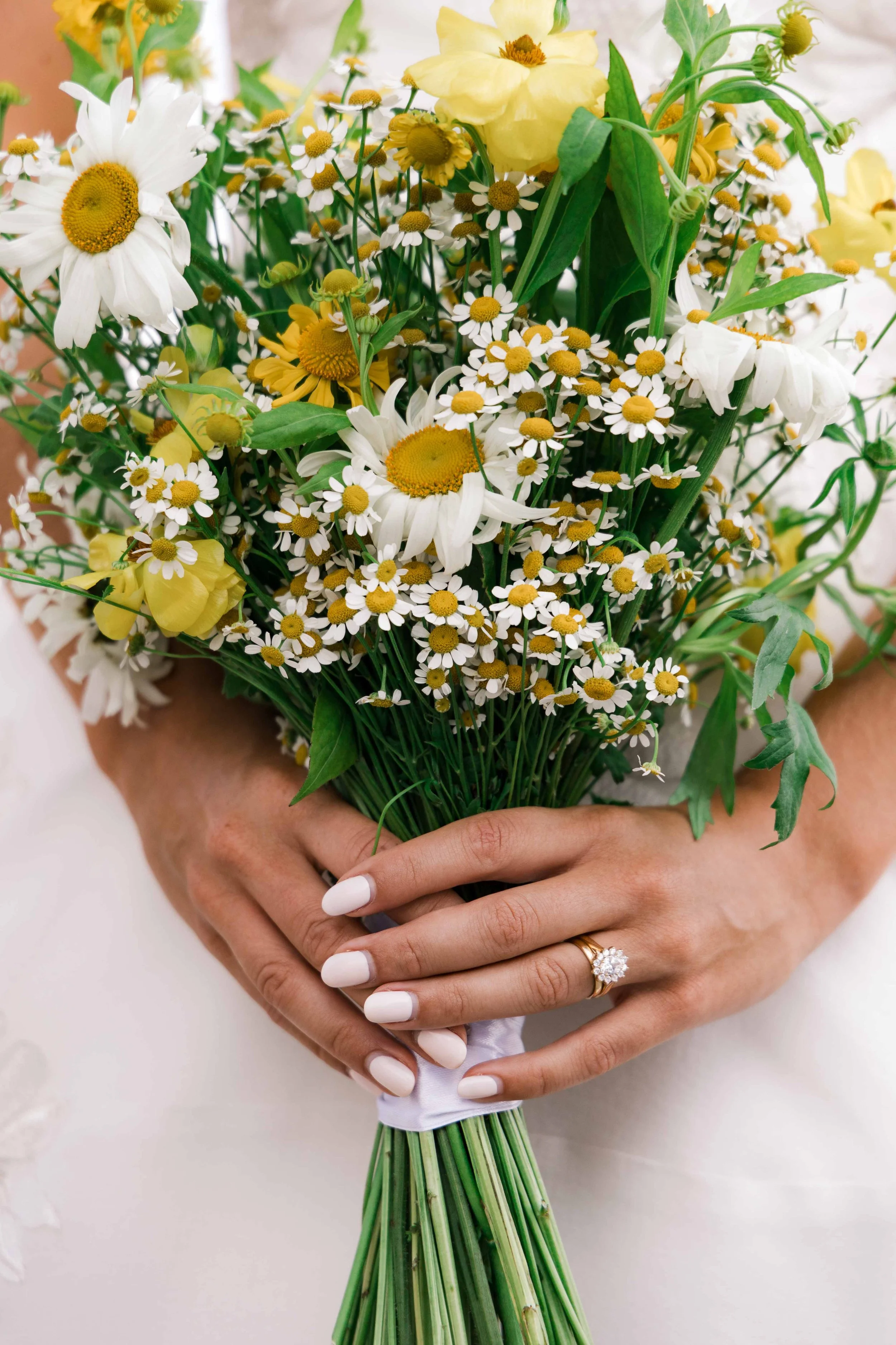 A woman is holding a bouquet of white daisies, yellow roses, and other yellow and white flowers, with her hands visible and adorned with a ring on her left ring finger. She has neatly manicured nails.