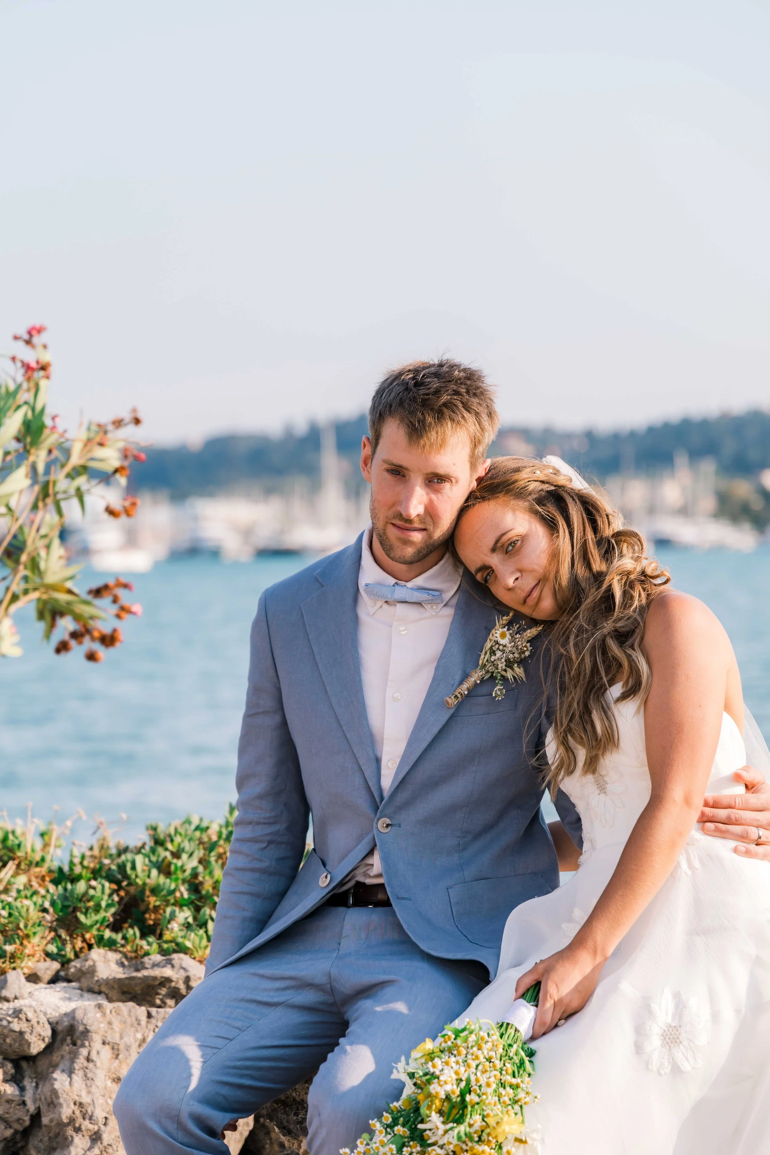 A couple dressed in wedding attire sitting by the water, with boats and hills in the background, during daytime.