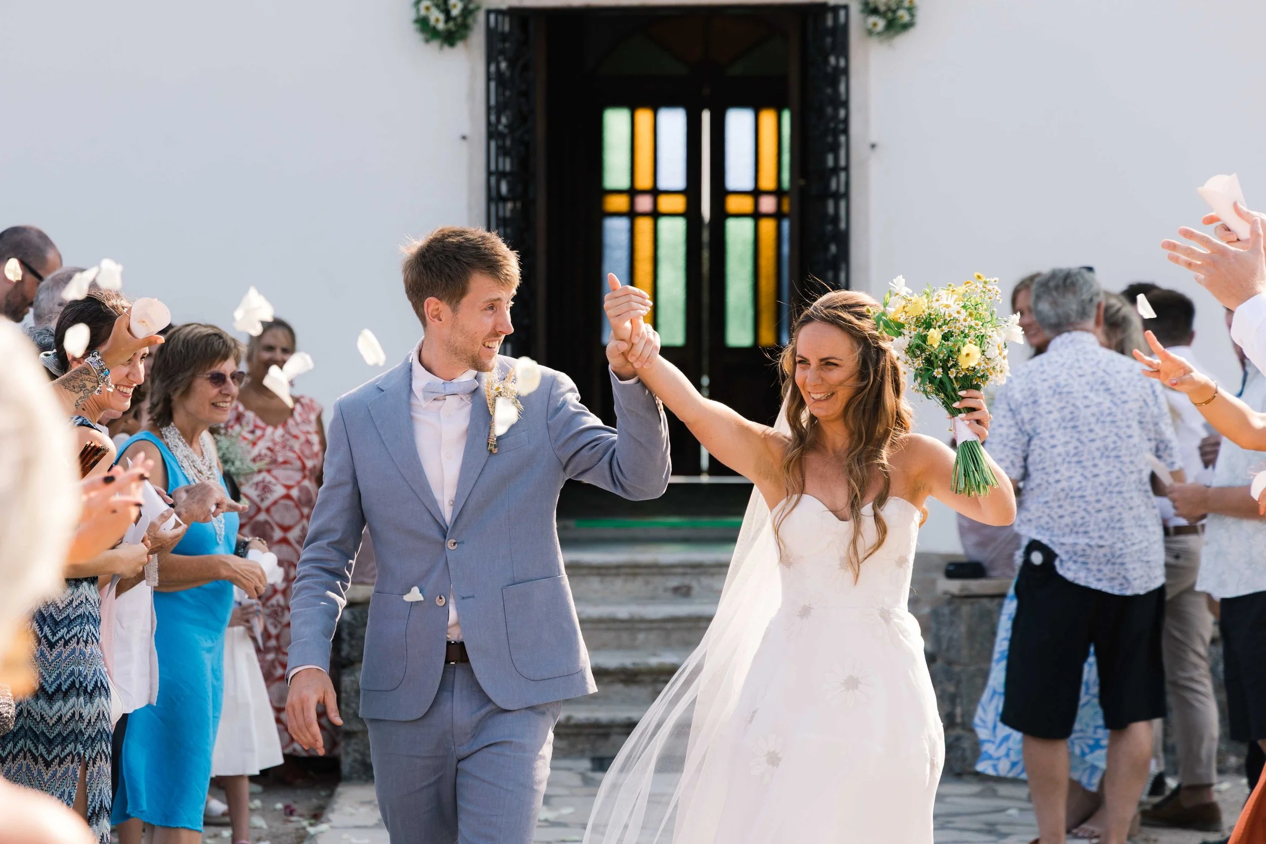 A bride and groom celebrating their wedding outside a church, surrounded by friends and family. The groom is wearing a light gray suit, and the bride is holding a bouquet of white and yellow flowers, smiling as they dance.