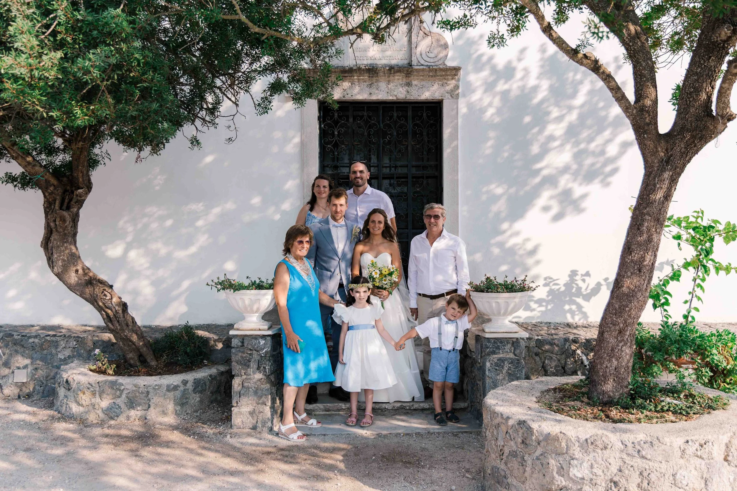 Group of people, including adults and children, posing outdoors in front of a white wall with a black gate, surrounded by trees and potted plants.