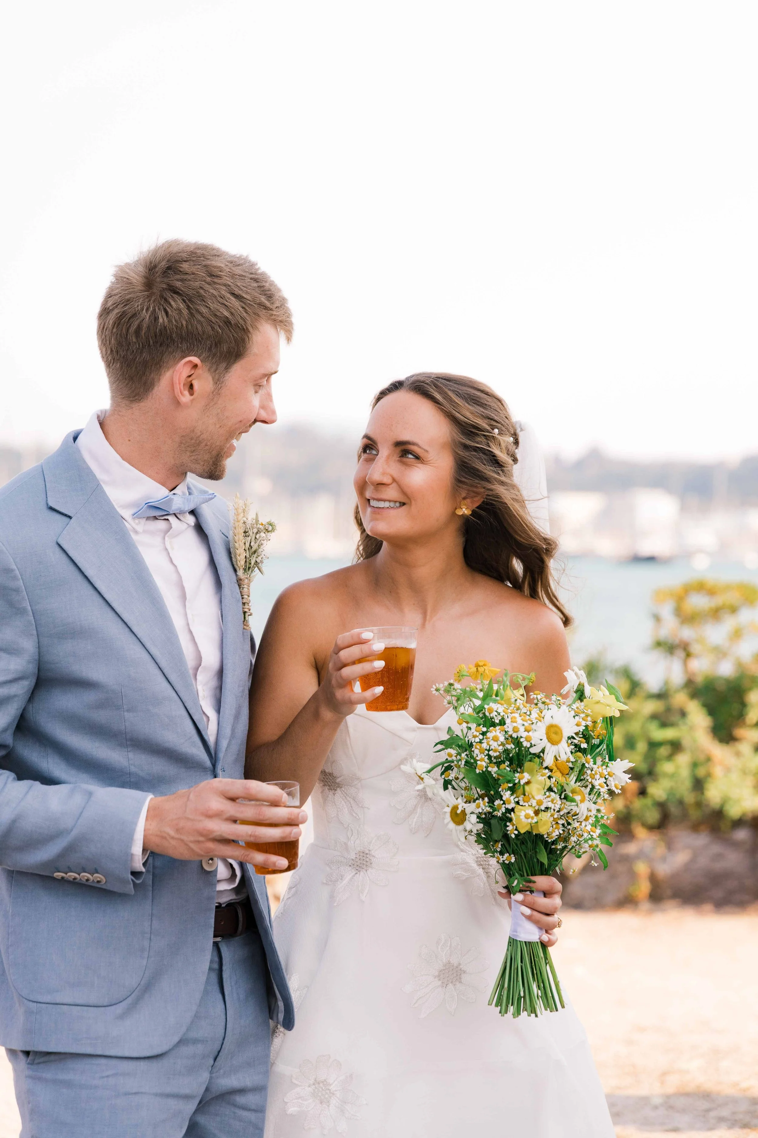 Bride and groom at their outdoor wedding, holding drinks and smiling at each other, with a scenic city view in the background.