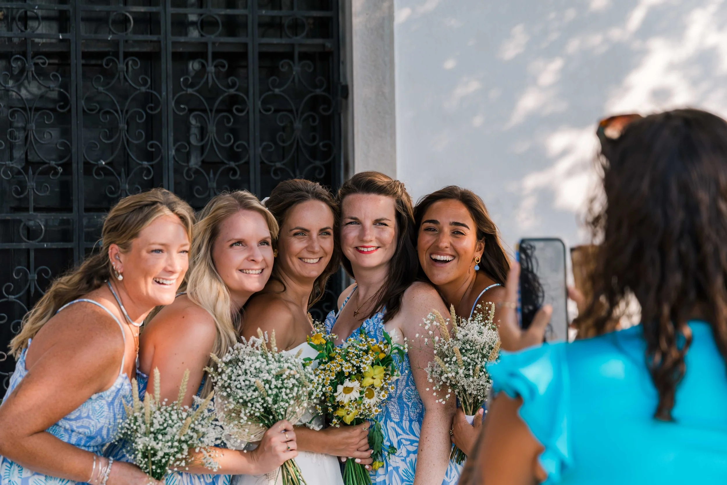 A group of women in blue and white dresses with floral patterns taking a selfie during a celebration, holding bouquets of flowers. They are smiling and posing outdoors near a dark ornate gate, with a partly cloudy sky in the background.