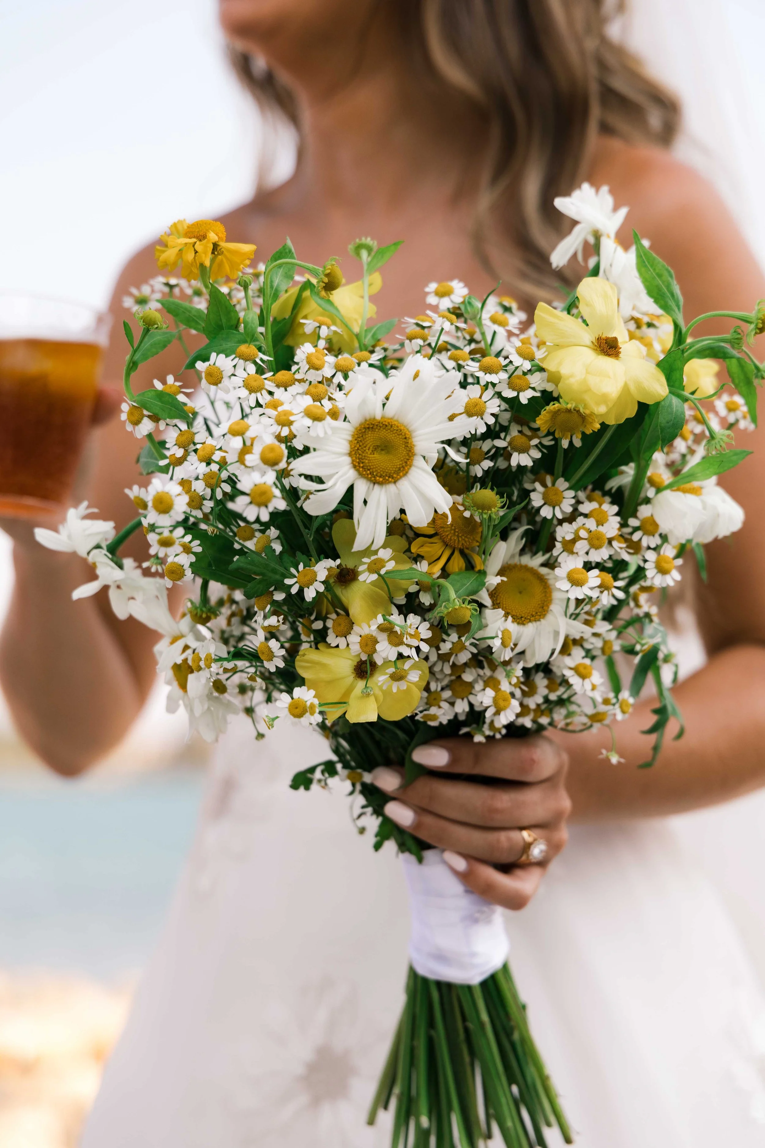 A woman in a white dress holding a bouquet of yellow and white flowers, with a blurred background.