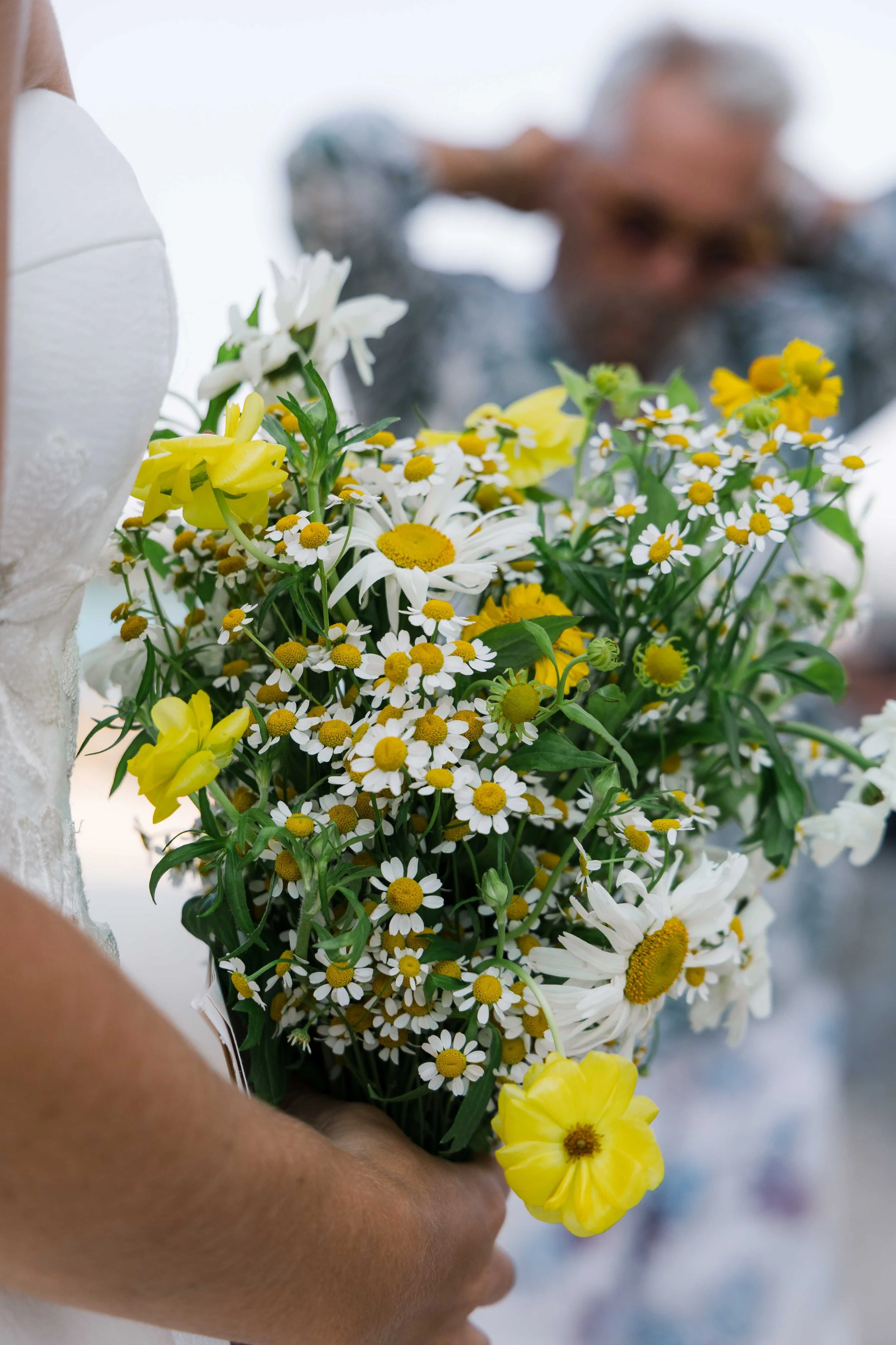 Close-up of a bouquet of yellow and white flowers being held by a person with a blurred background of a woman with gray hair and glasses.