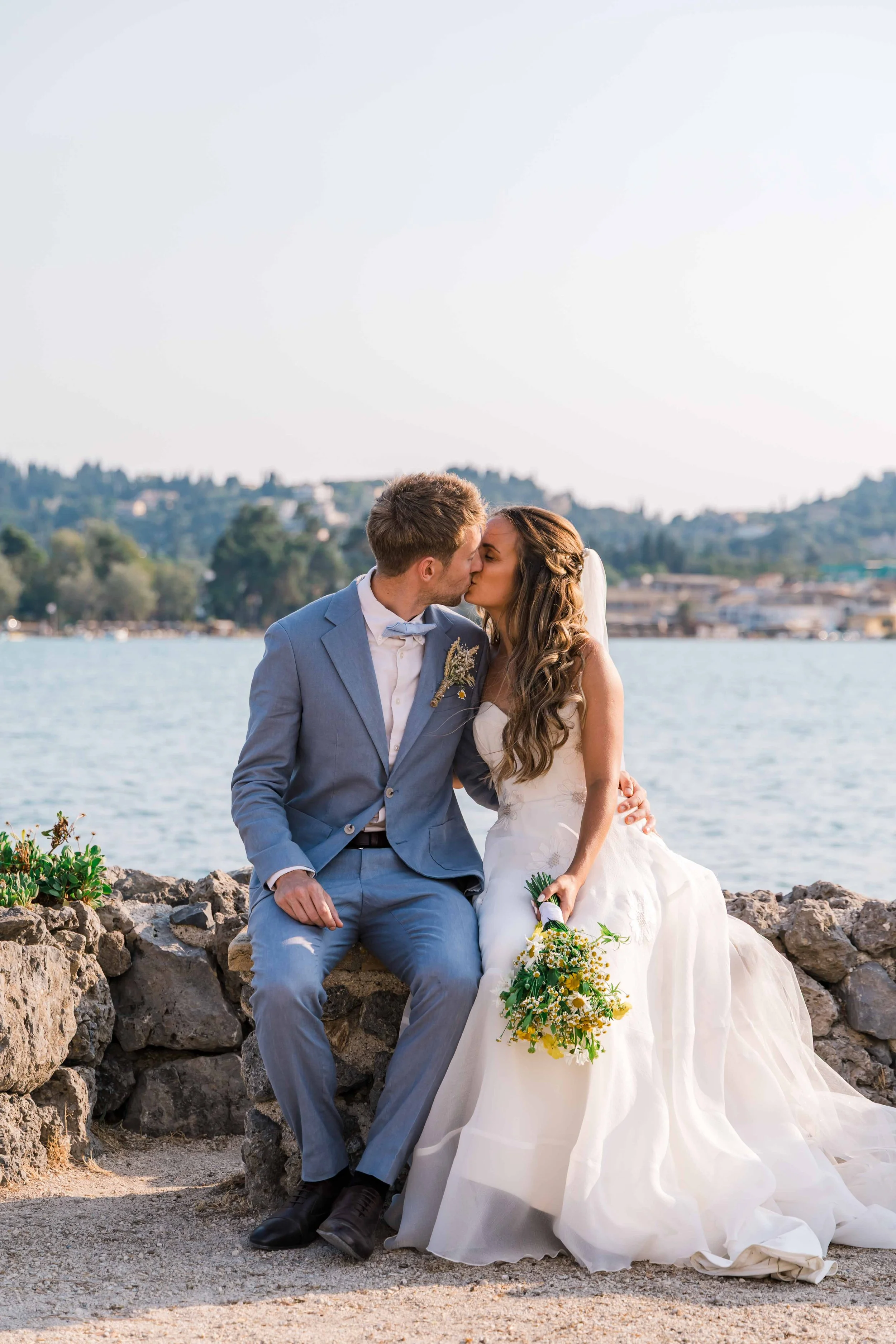 A bride and groom sitting on rocks near a body of water, kissing, with city and hills in the background. The bride wears a white wedding gown and holds a bouquet, while the groom wears a gray suit and white shirt.