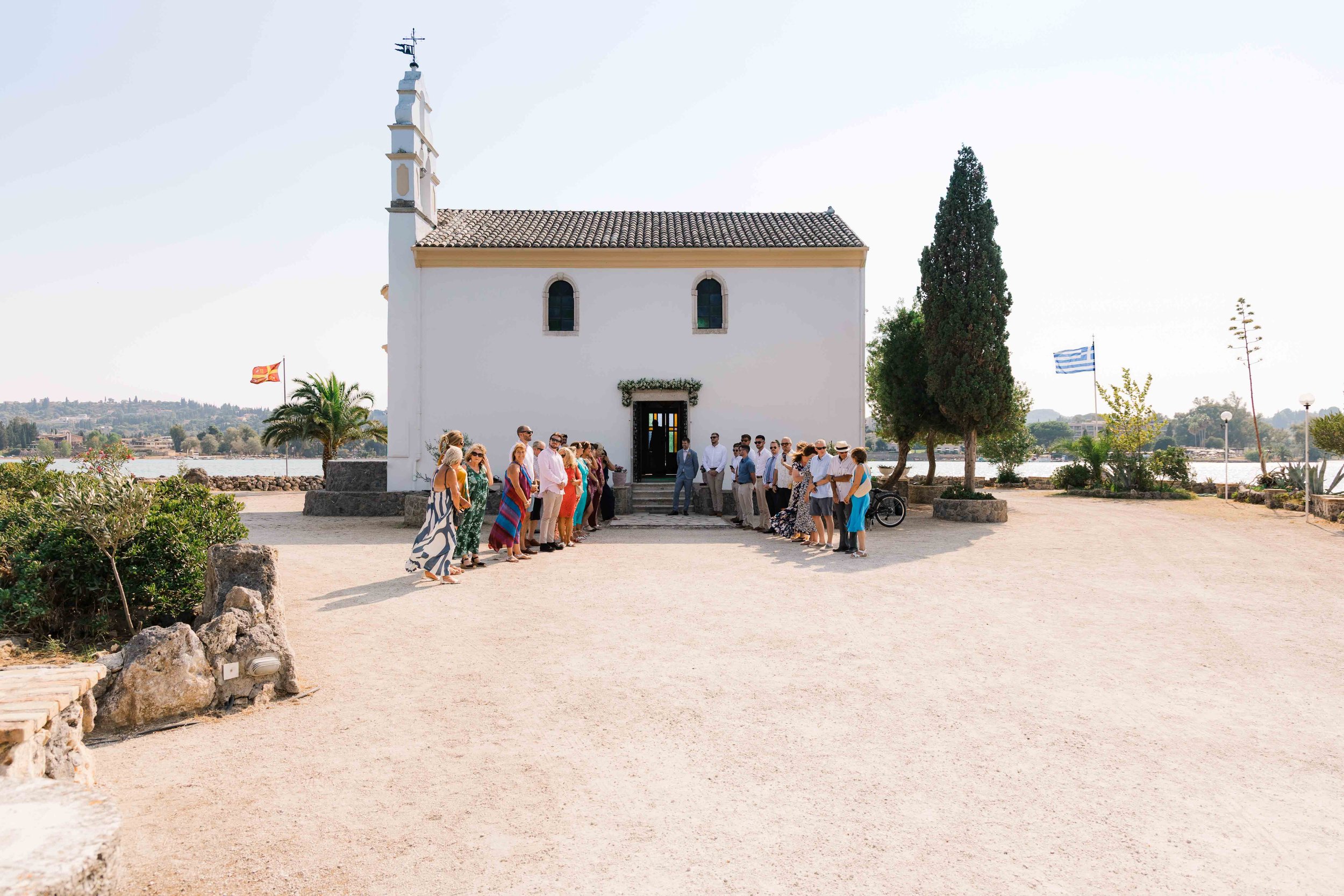 A group of people dressed in colorful summer clothing standing outside a white church with arched windows and a tiled roof. The church is near a body of water, with trees and flags, including a Greek flag, in the background.
