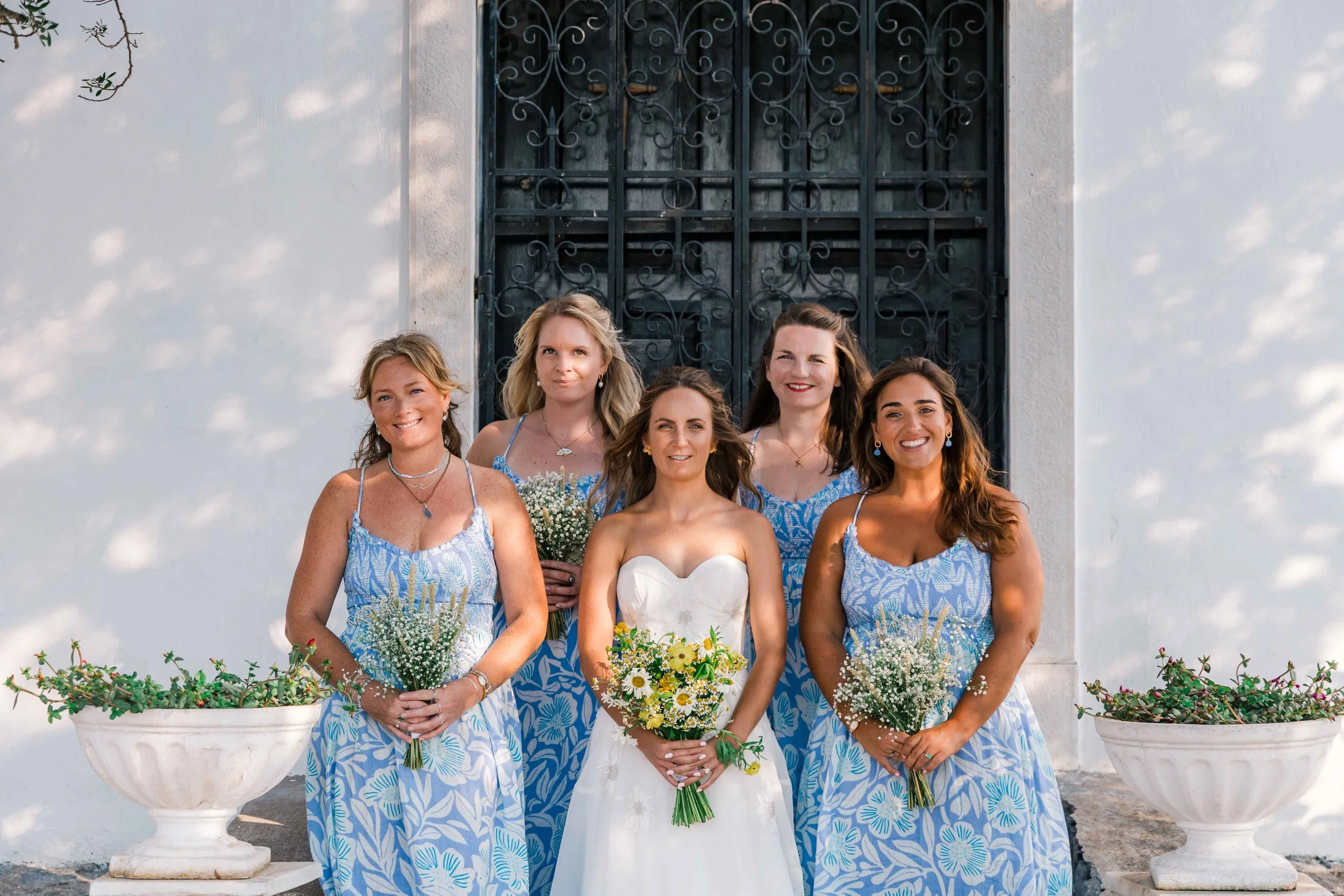 Six women standing outdoors in front of a white wall with ornate black window bars, four wearing matching blue and white patterned dresses, with the bride in the center wearing a white dress, all holding bouquets of flowers, smiling for a group photo