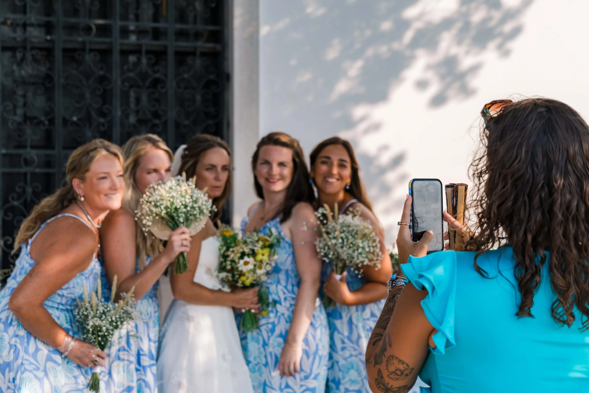 A group of women at a wedding, some holding bouquets of flowers, taking a photo together outdoors near a building with decorative black metalwork on the door, in sunny weather.