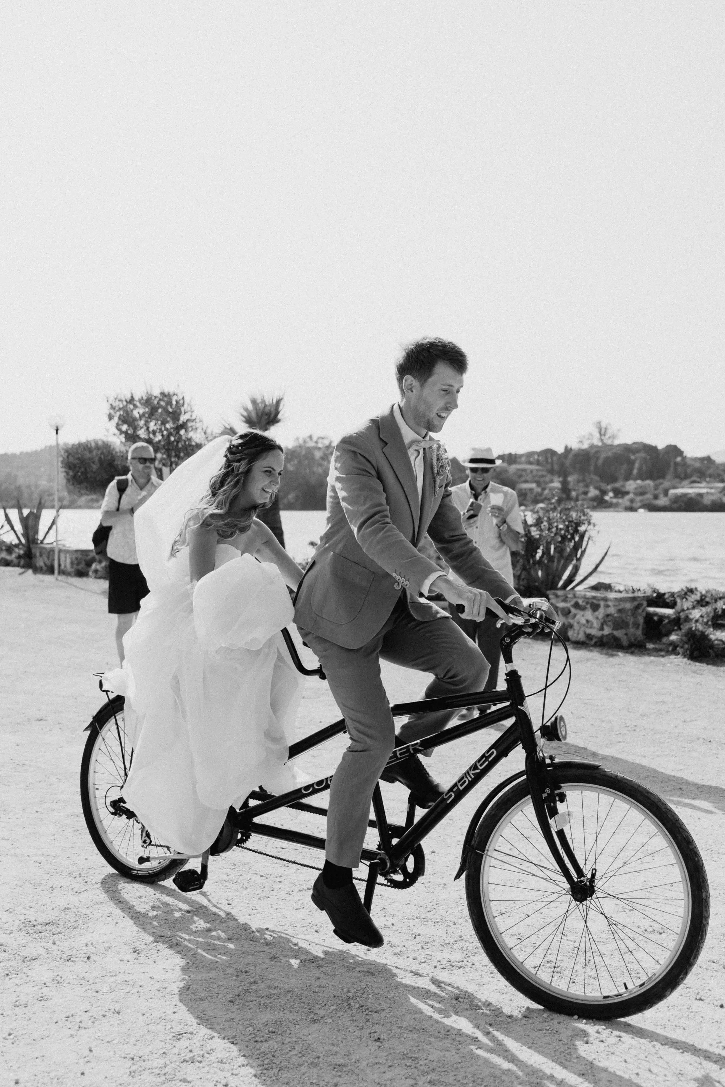 A man riding a bicycle with a wedding dress and veil on the back, smiling, with a woman in the background taking a photo, outdoors near water, with trees and hills in the distance.