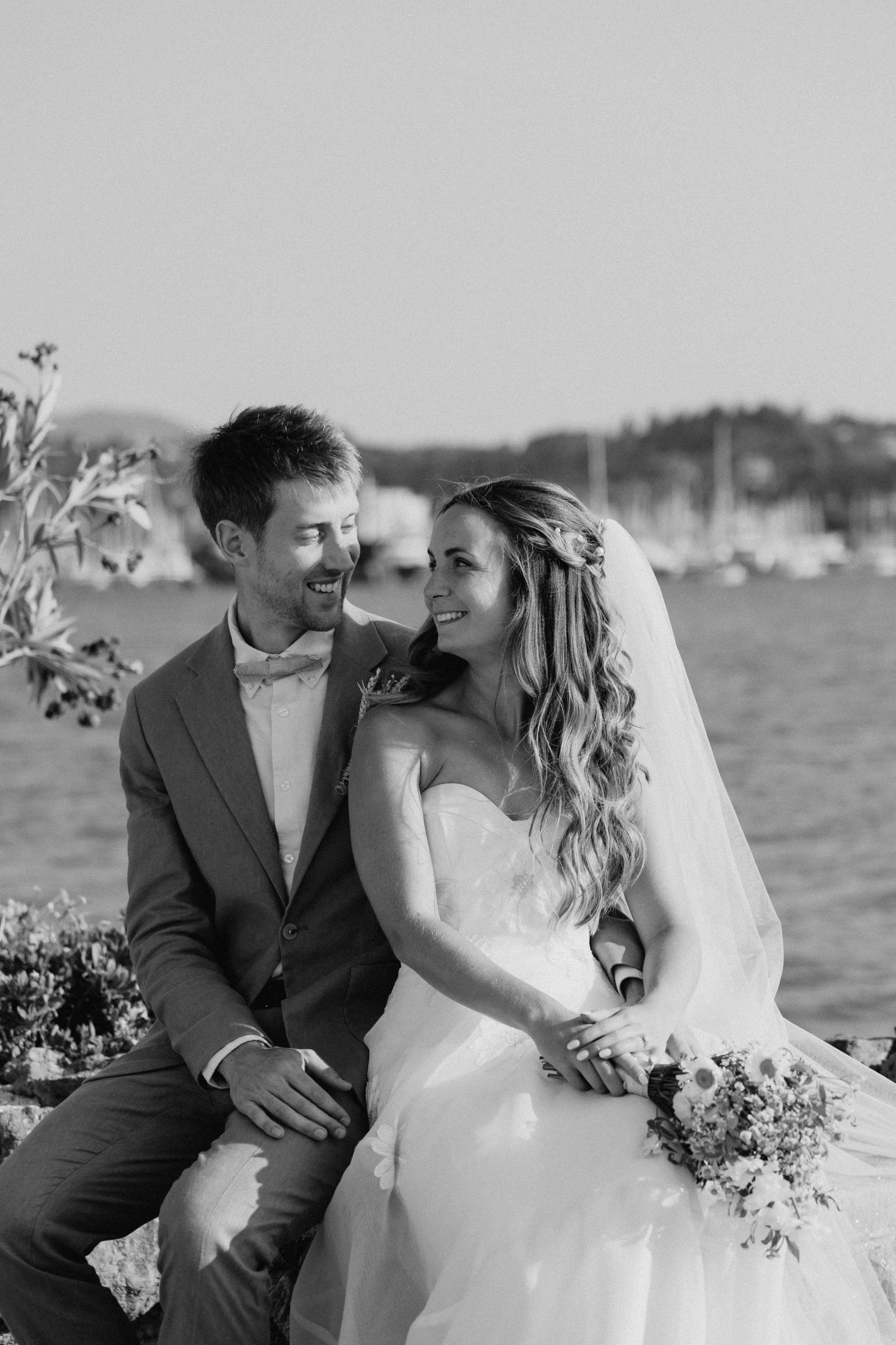 A black and white photo of a smiling man and woman in wedding attire sitting outdoors near a body of water with boats and a distant landscape in the background. The woman is wearing a wedding dress and veil, holding a bouquet of flowers, and looking 
