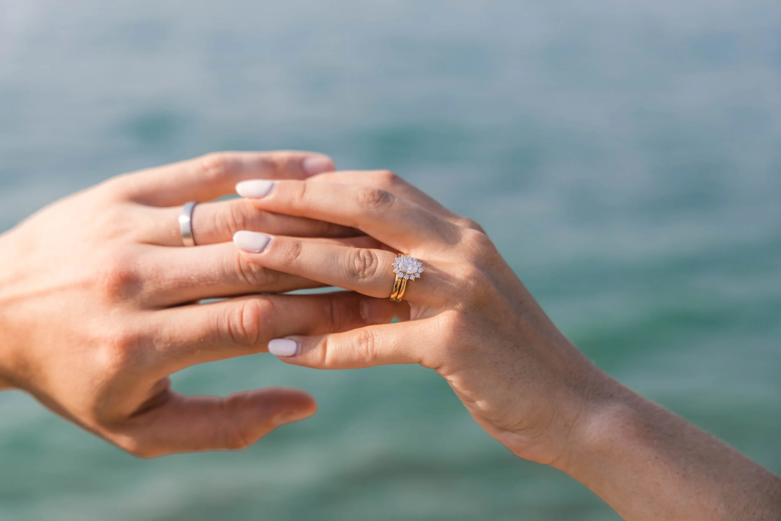 Close-up of two hands holding each other, with one wearing a silver ring and the other wearing a gold ring with a cluster of diamonds, against a backdrop of a blue ocean.
