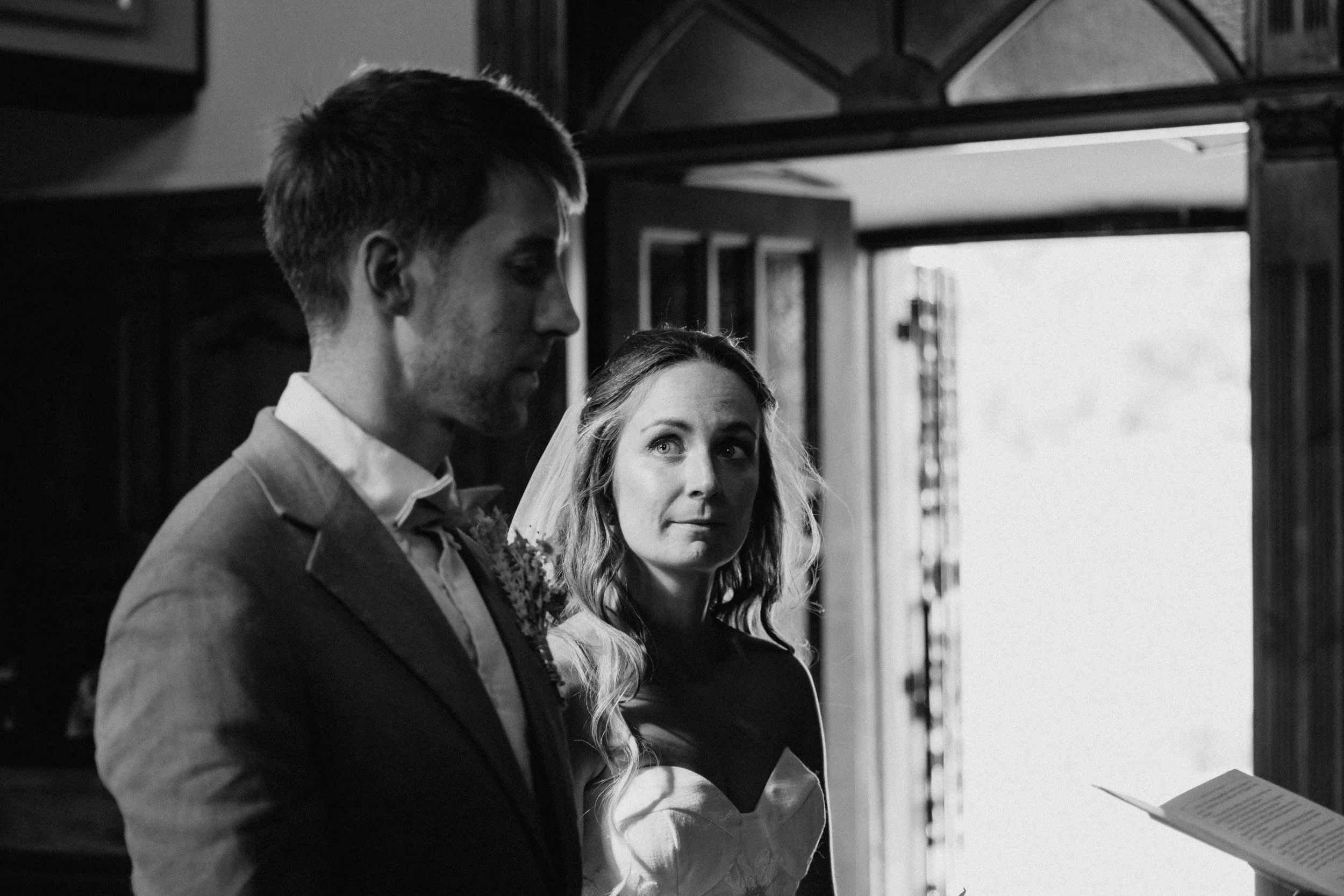 A black and white photo of a man in a suit and a woman with long hair, both looking at a book or paper, inside a room with a door and window in the background.