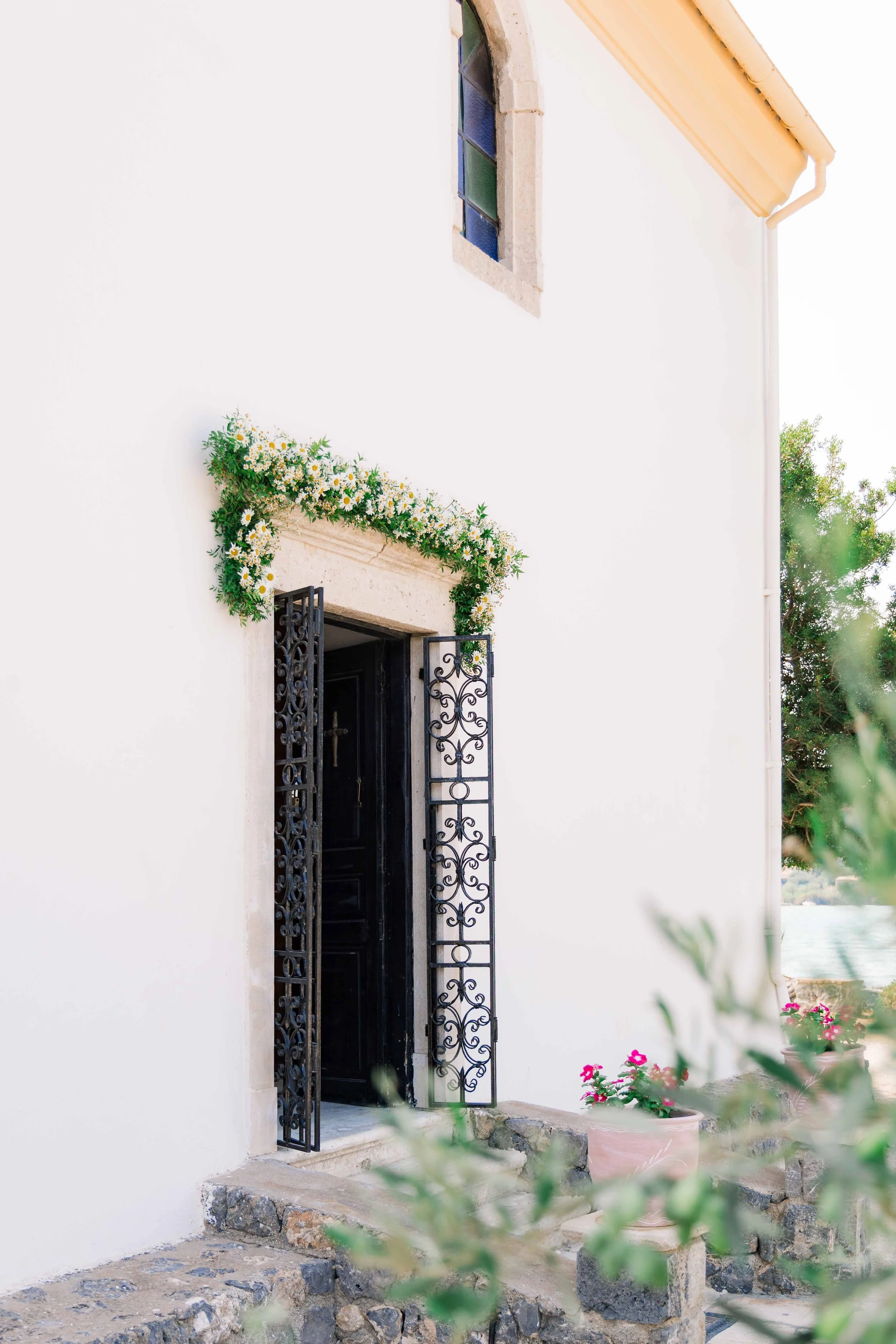 Exterior view of a white building with a small arched stained-glass window. An open decorative black iron gate is beside the entrance, which is surrounded by a floral border with white and yellow daisies. Potted pink flowers and green foliage are nea
