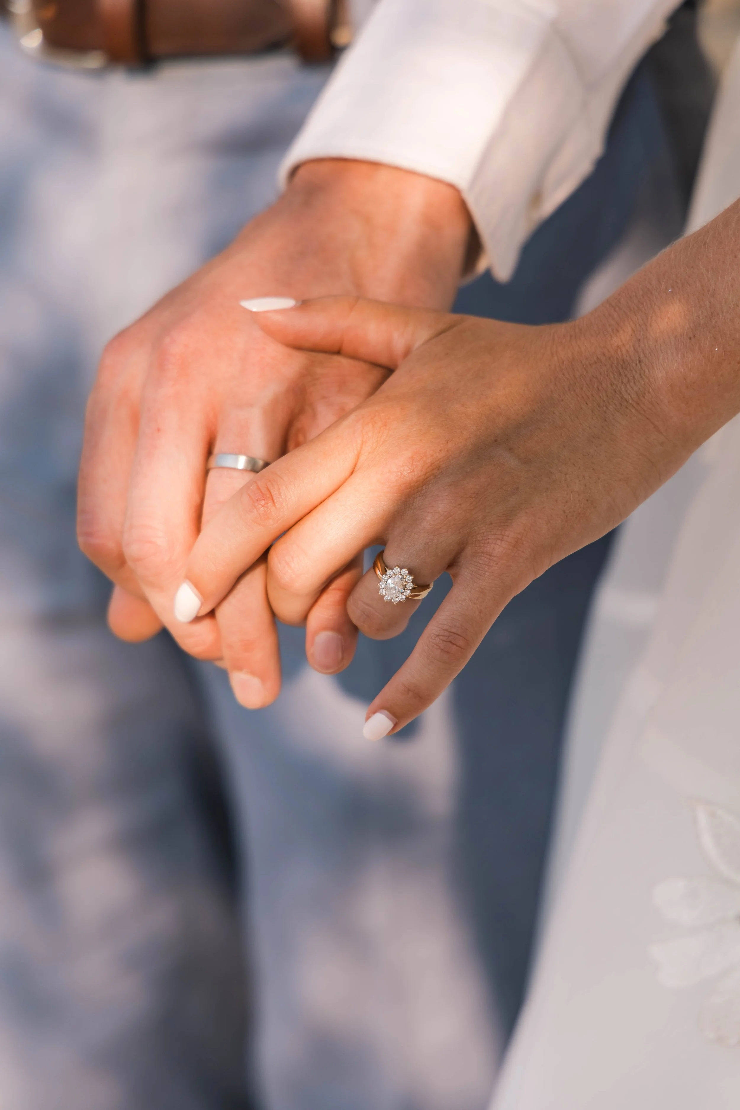 Close-up of a couple holding hands, both wearing wedding rings, with the woman wearing a ring with a large floral design. One hand appears to be slightly darker in skin tone than the other.