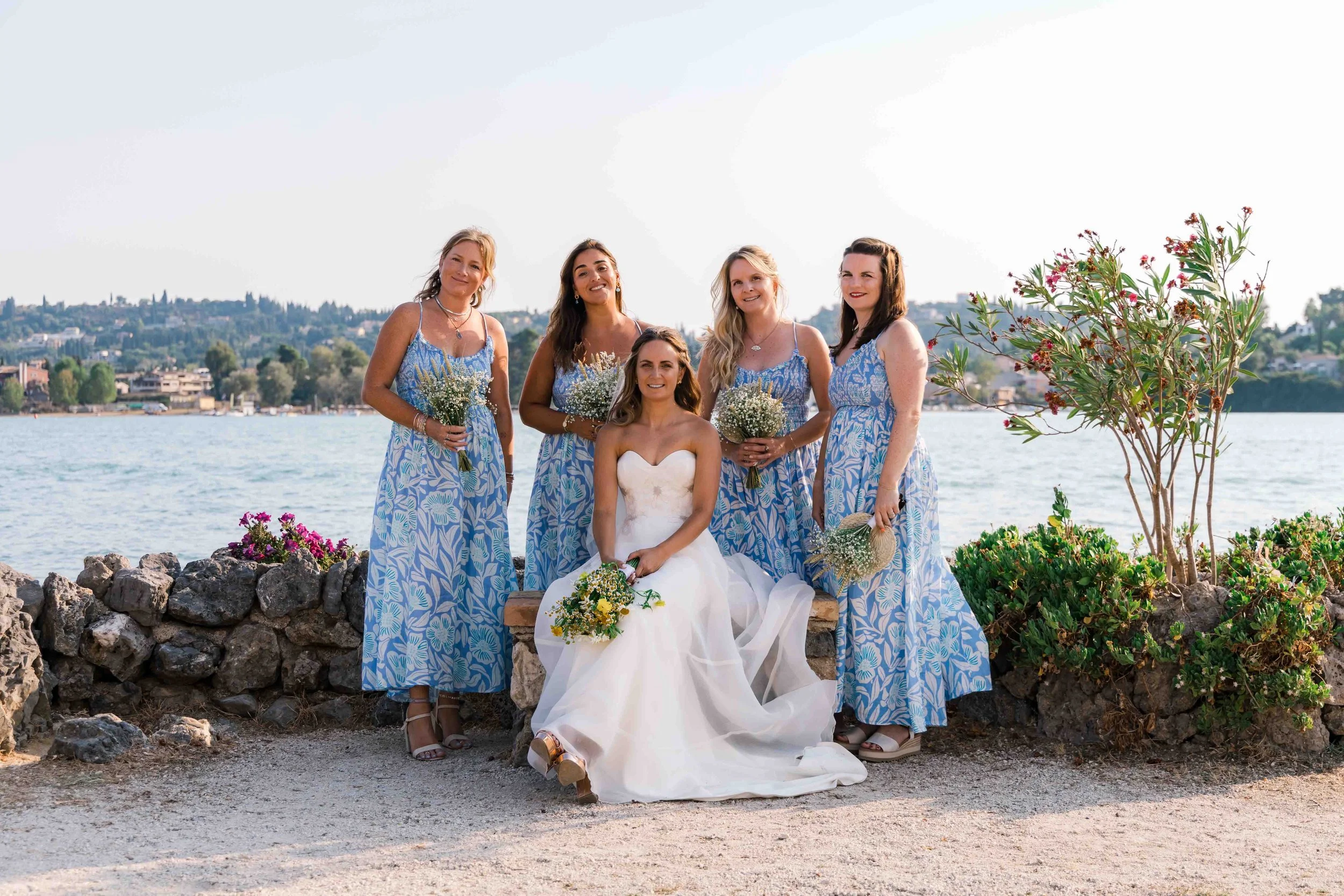A group of six women, one in a white wedding dress and the others in matching blue and white patterned dresses, pose outdoors near a body of water with hills and houses in the background. One woman is seated while the others stand behind her, all smi