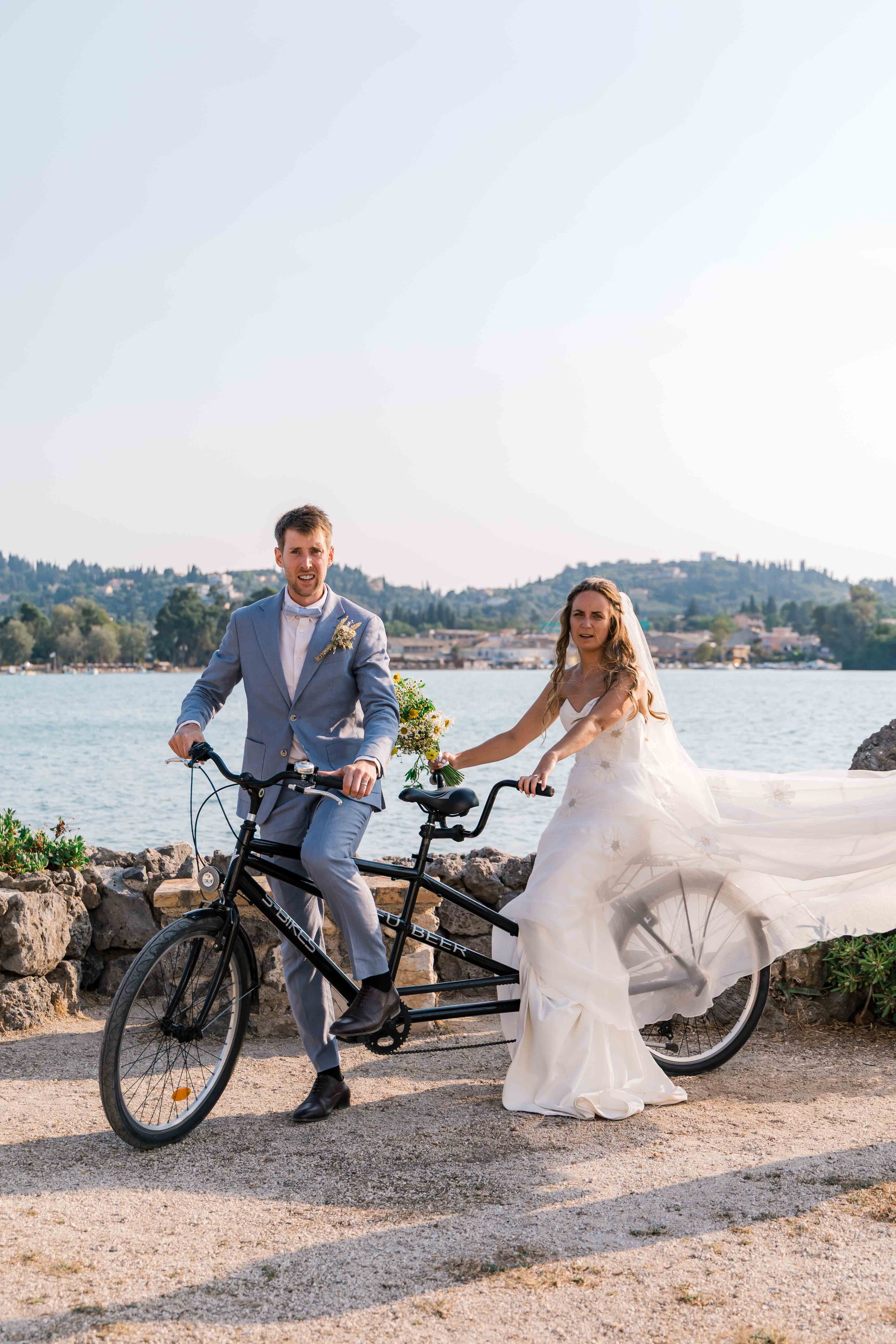 A newlywed couple is riding a tandem bicycle along a waterfront path, with the groom in a gray suit and the bride in a white wedding dress, holding a bouquet, and wearing a veil, with hills and water in the background.