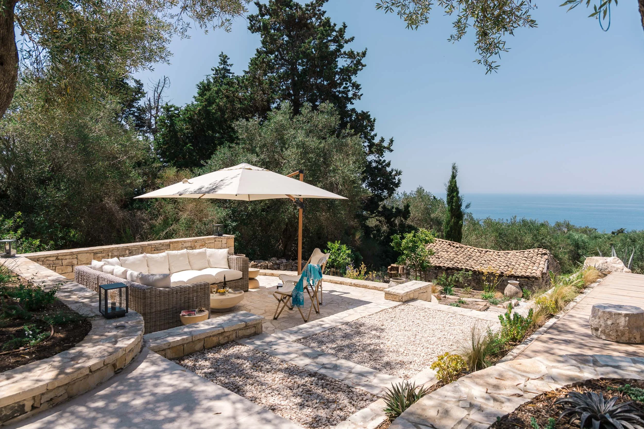 A serene outdoor patio with a cushioned wicker sofa under a large white umbrella, surrounded by lush green trees, overlooking a view of the ocean in the distance.