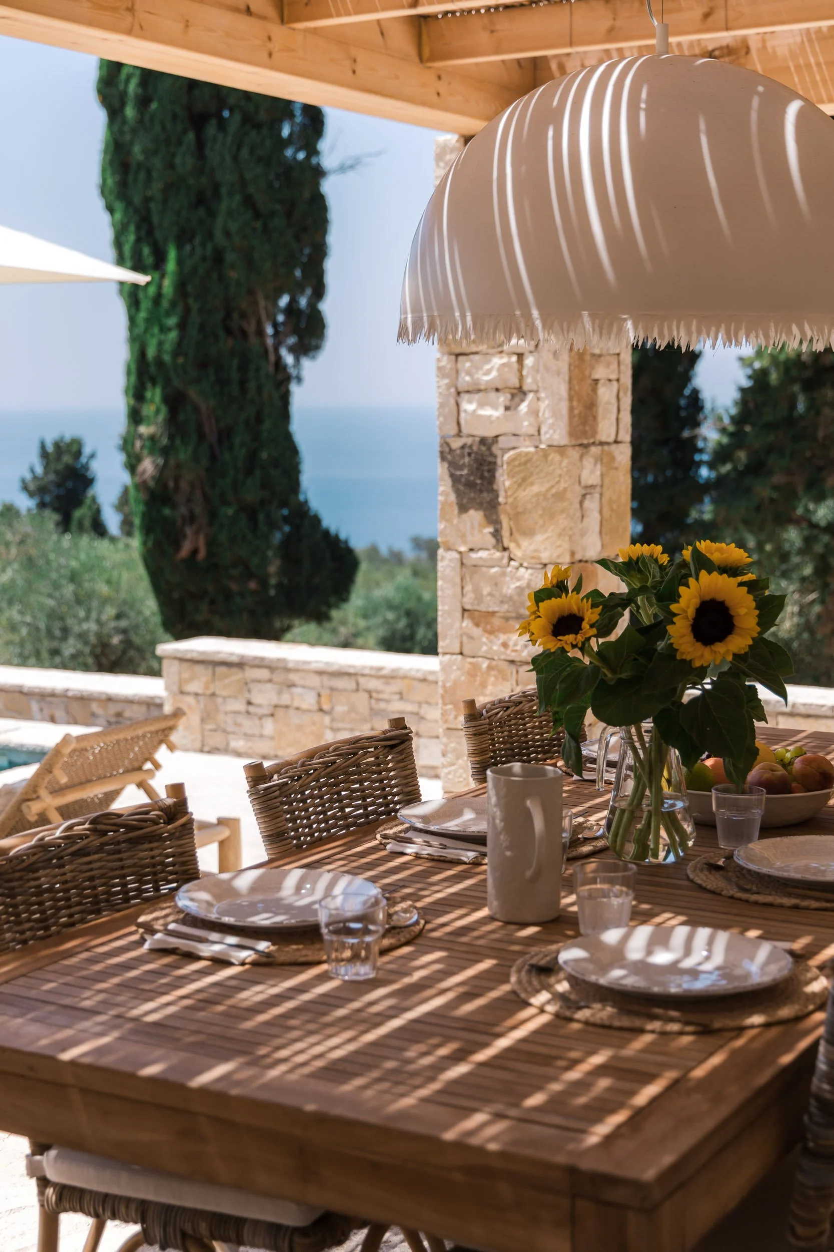 Outdoor dining table with sunflower centerpiece, plates, mugs, and glasses, shaded by an umbrella, with stone wall and trees in the background overlooking the ocean.