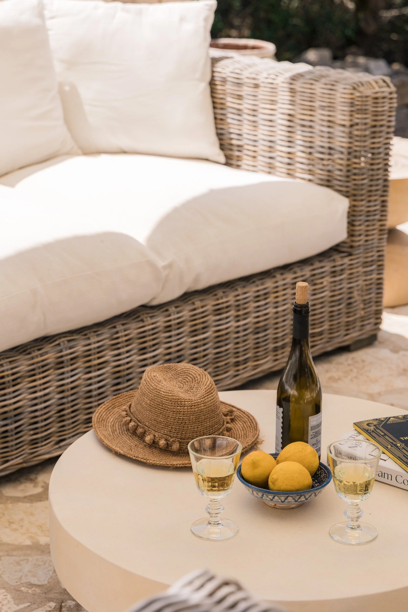 A wicker outdoor sofa with white cushions, a round white patio table with a straw hat, three glasses of white wine, a bowl of four yellow lemons, a bottle of wine with a cork, and a book.