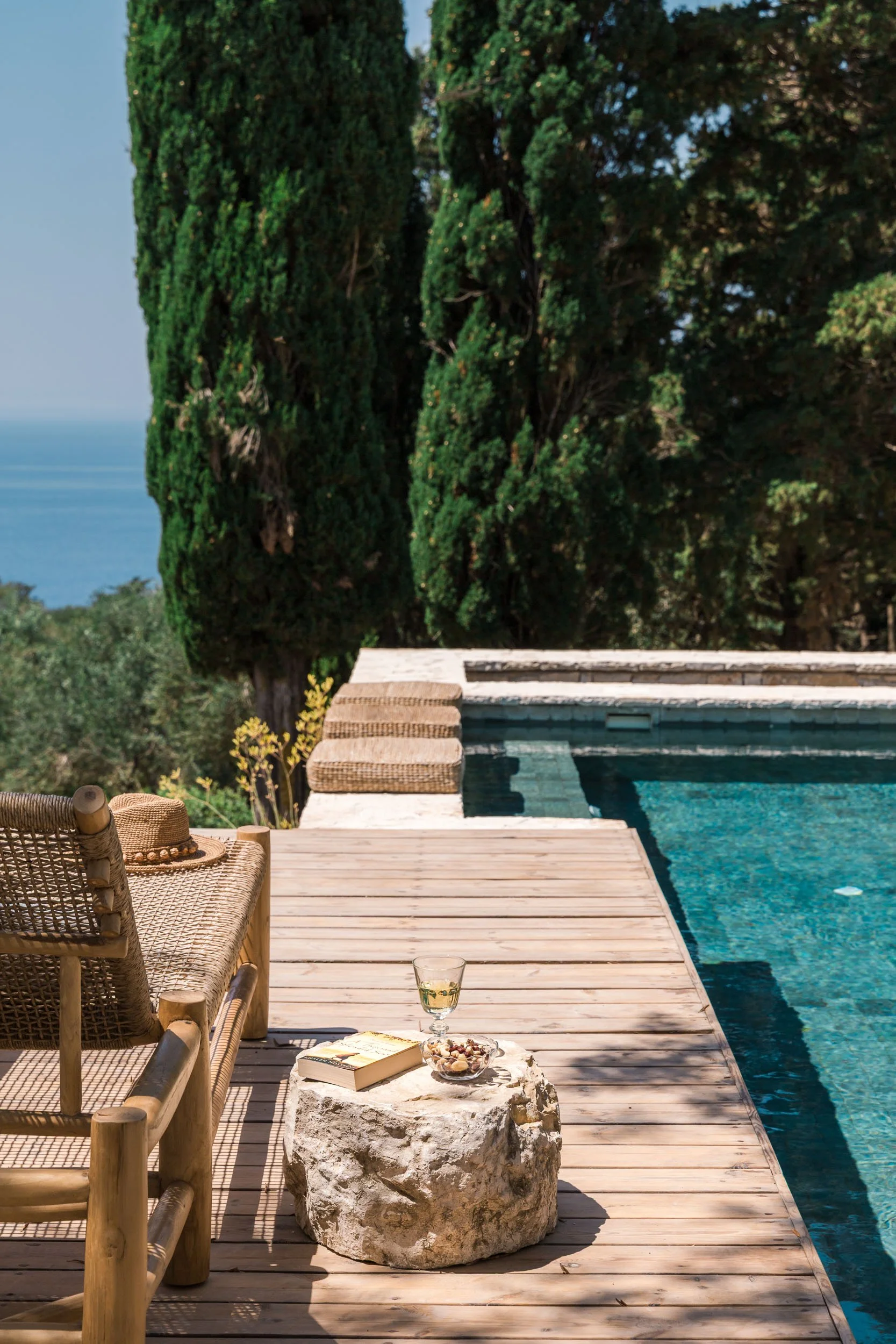 A wooden deck beside a swimming pool with a rocky side, surrounded by green trees. A chair with a hat, a glass of white wine, a book, and a bowl of snacks are on a rock table near the pool.