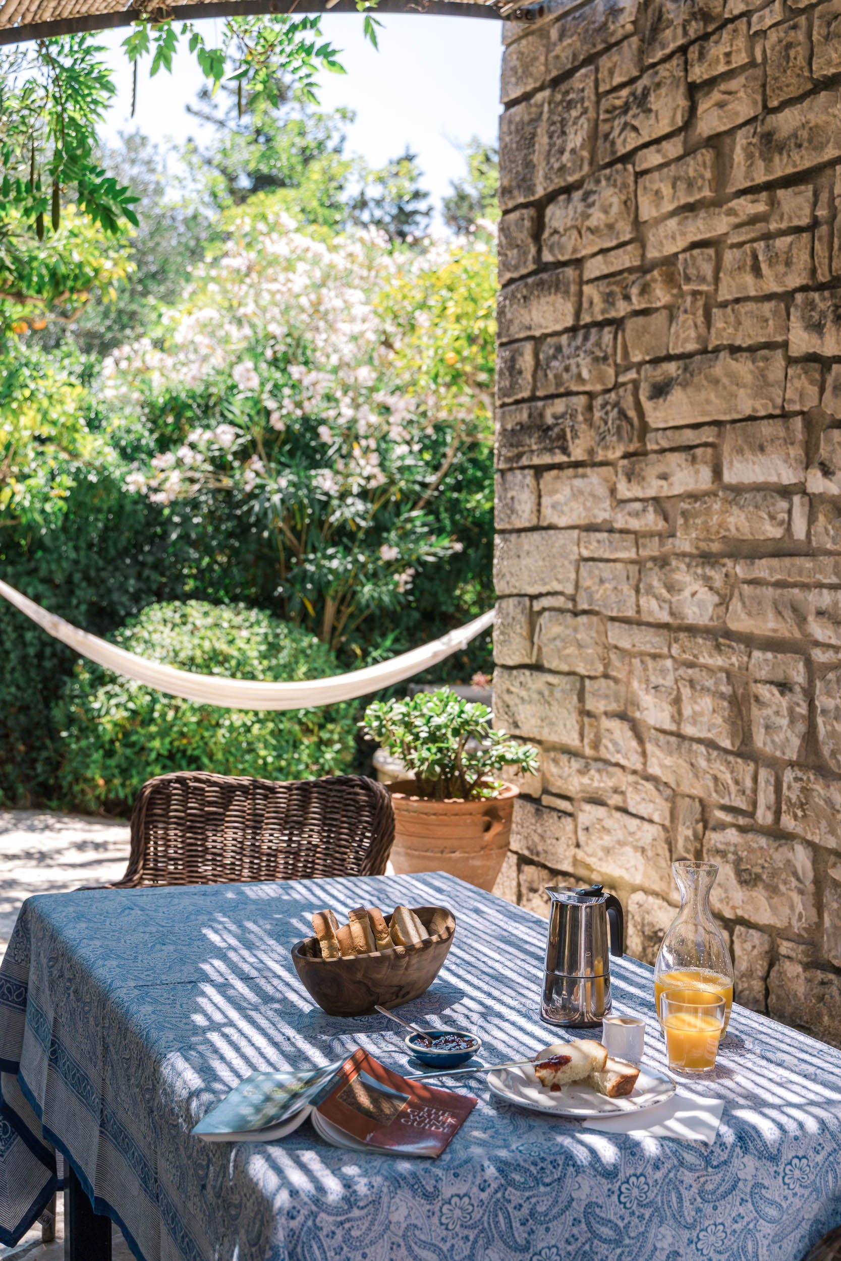 A table set for breakfast or brunch on an outdoor patio with a stone wall, surrounded by lush greenery and pink flowering bushes. The table has a blue patterned tablecloth, a bowl of bread, a plate with a half-eaten pastry, a glass pitcher of orange 