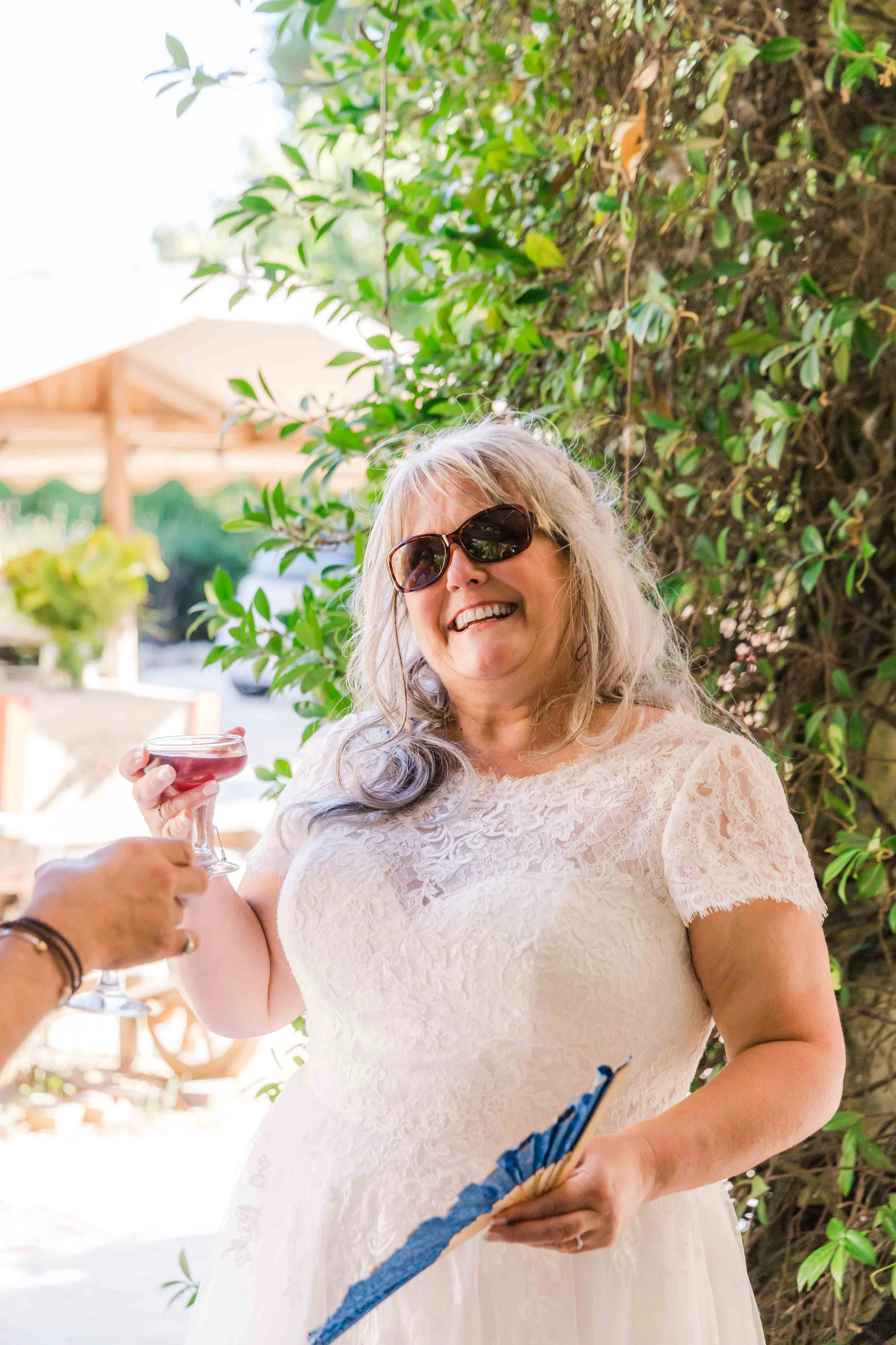A woman in a white lace dress and sunglasses smiling as she receives a toast of red wine from another person at an outdoor gathering with greenery and a wooden structure in the background.