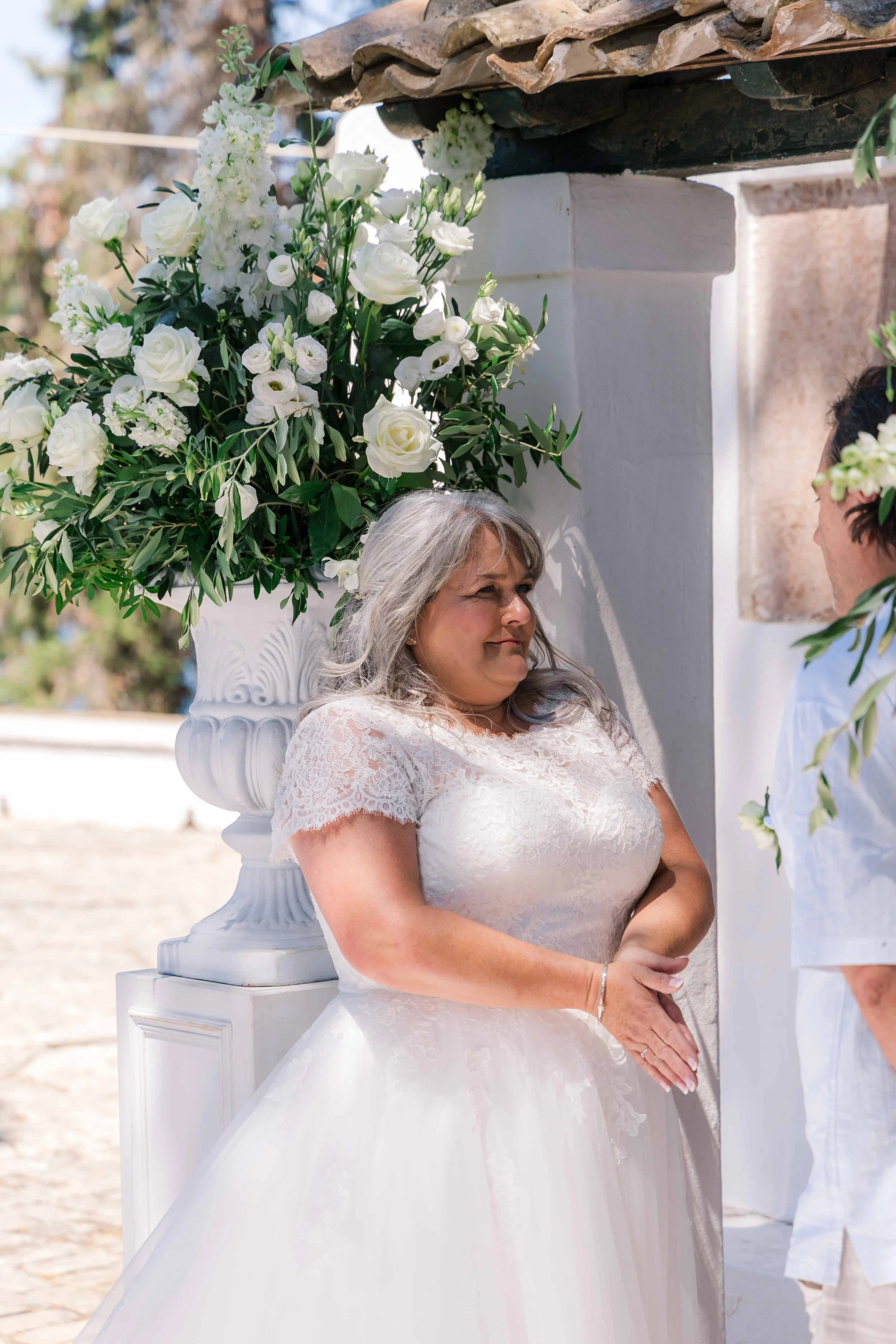 A woman dressed in a white wedding gown standing beside a large floral arrangement with white roses and greenery during a wedding ceremony outside.