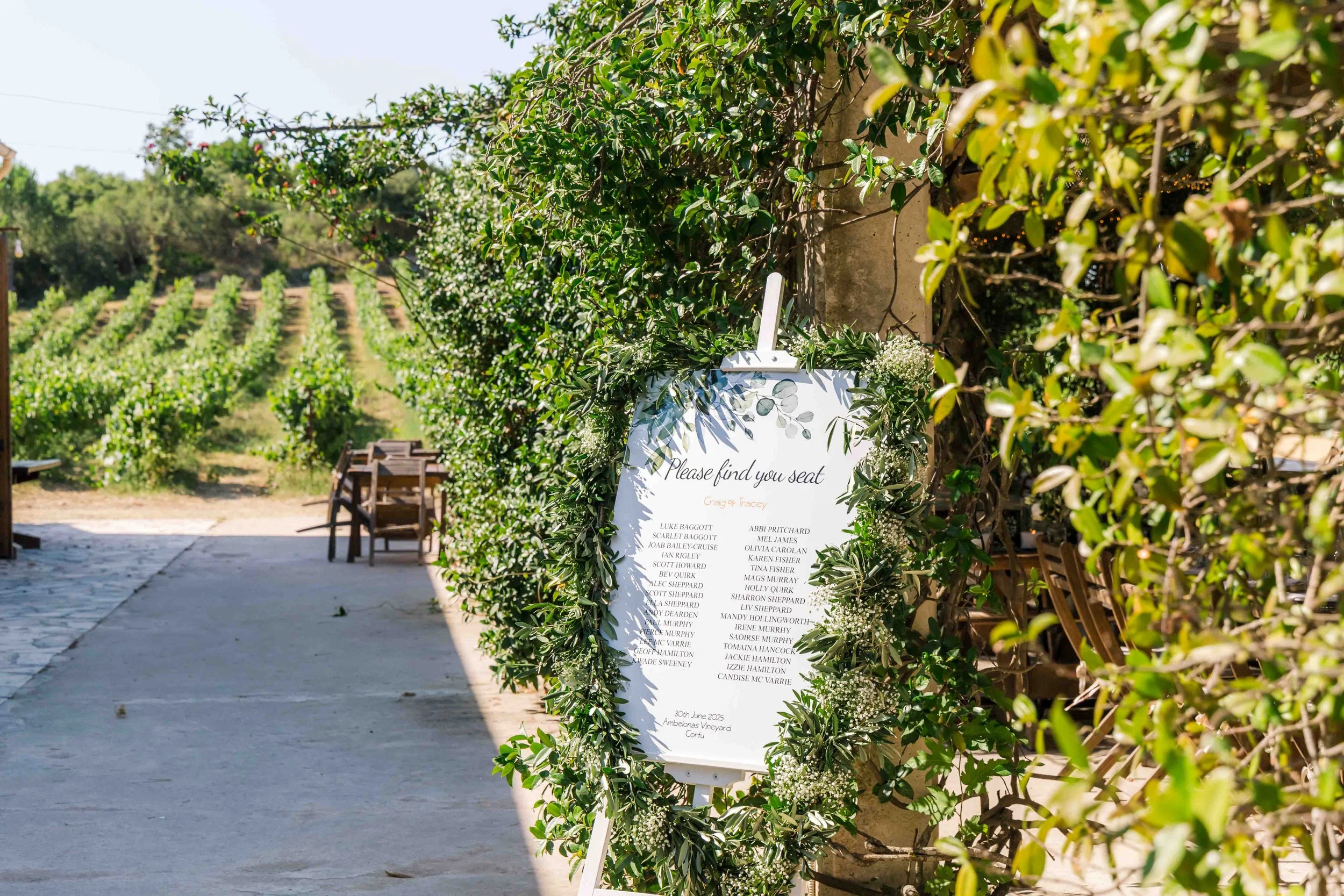 Wedding seating chart with greenery decorations at an outdoor venue, with chairs and a vineyard in the background.