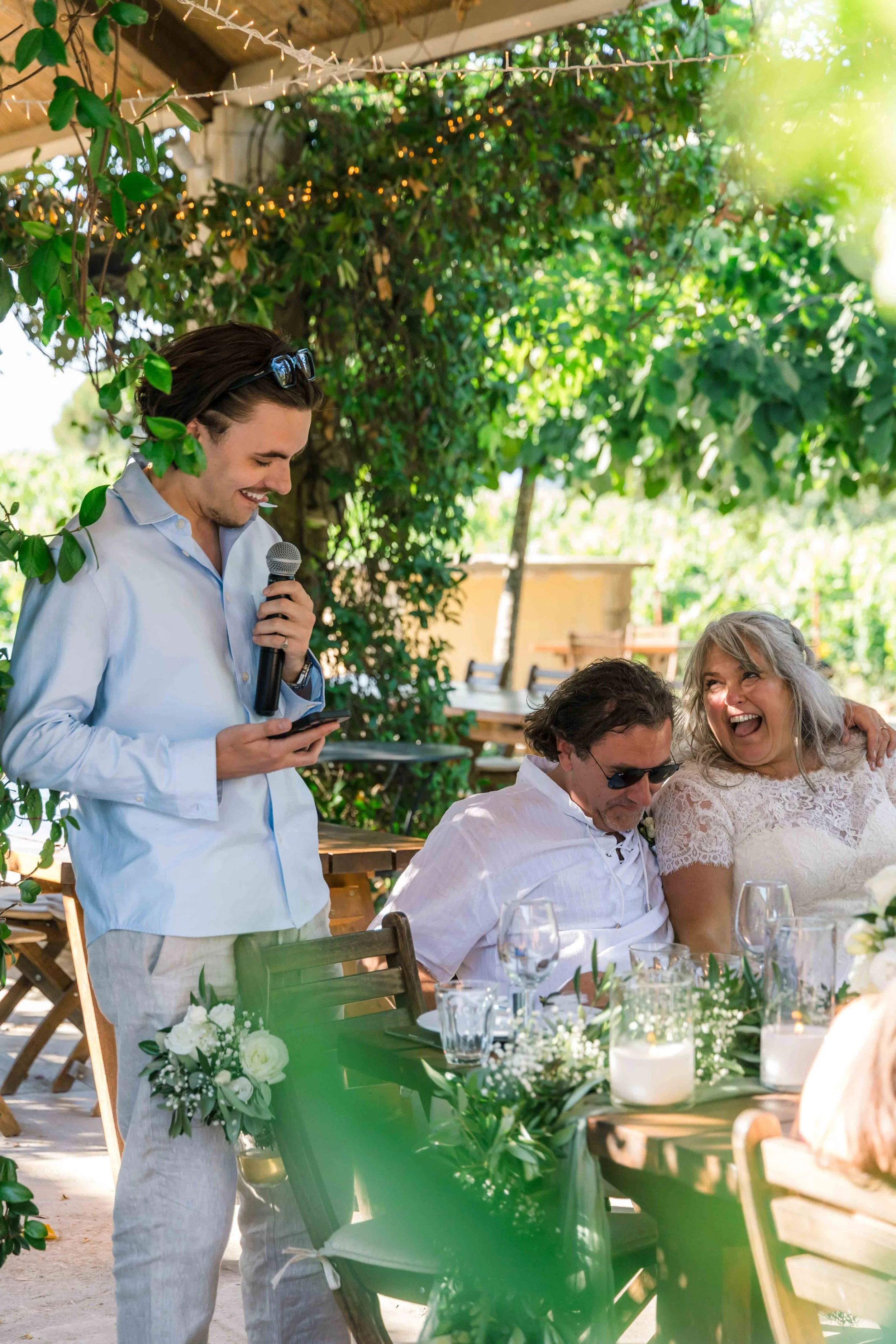 A young man in a light blue shirt and beige pants, holding a microphone and a phone, is speaking at an outdoor gathering. Three other people, two men and a woman, are seated at a table decorated with flowers and candles, smiling and engaging in conve