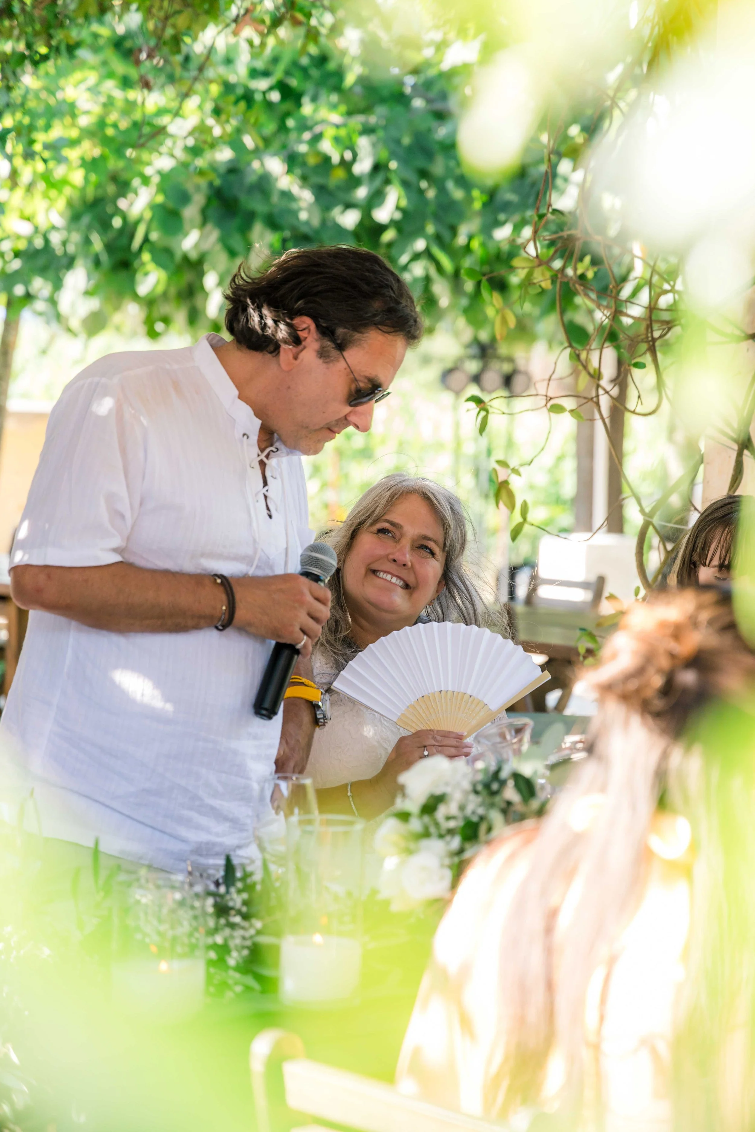 A man in a white shirt and sunglasses holding a microphone, sitting at a table outdoors, smiling at a woman holding a fan, with greenery and sunlight in the background.