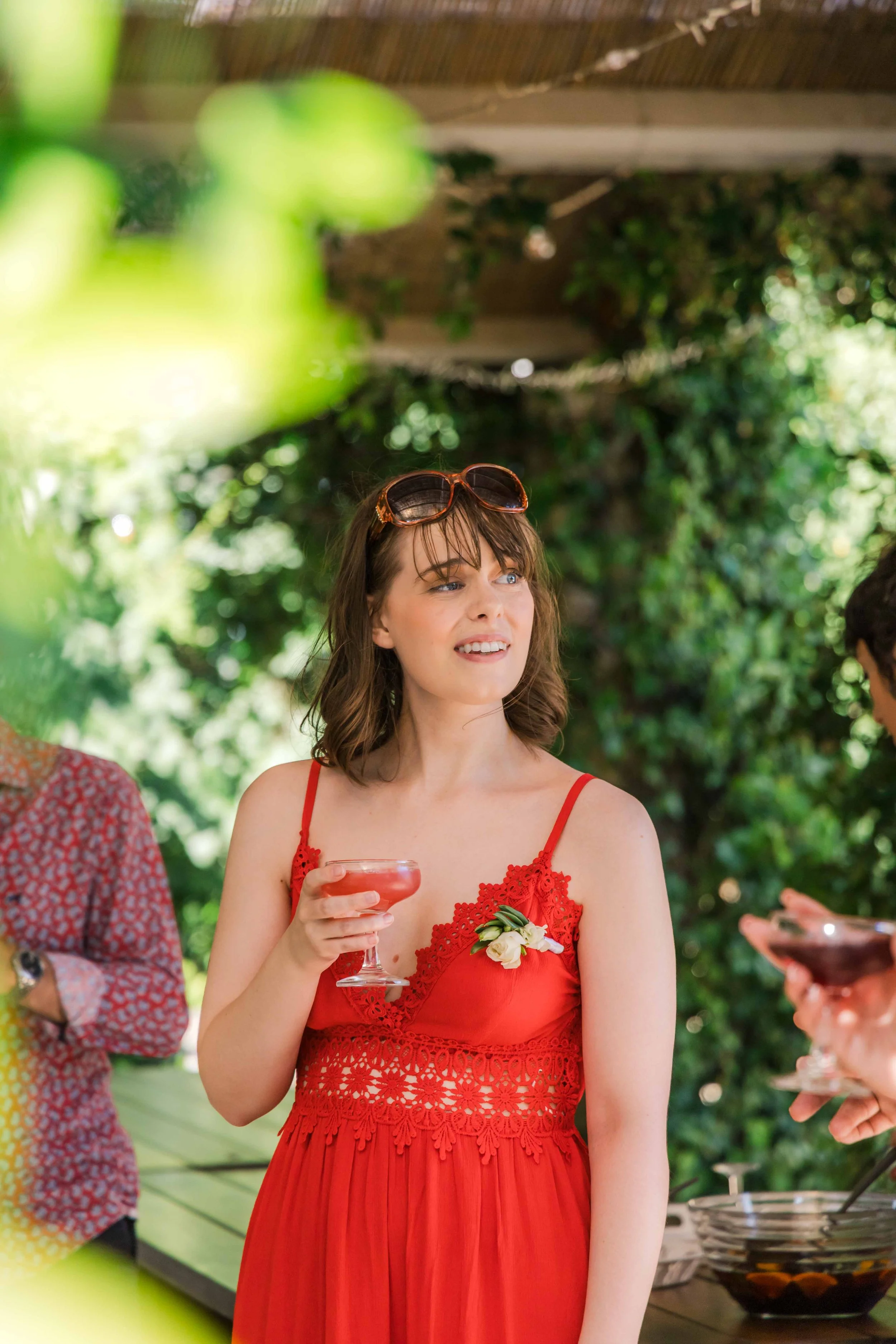 A woman with shoulder-length brown hair, wearing a red dress and sunglasses on her head, holding a cocktail glass, at an outdoor gathering surrounded by greenery.